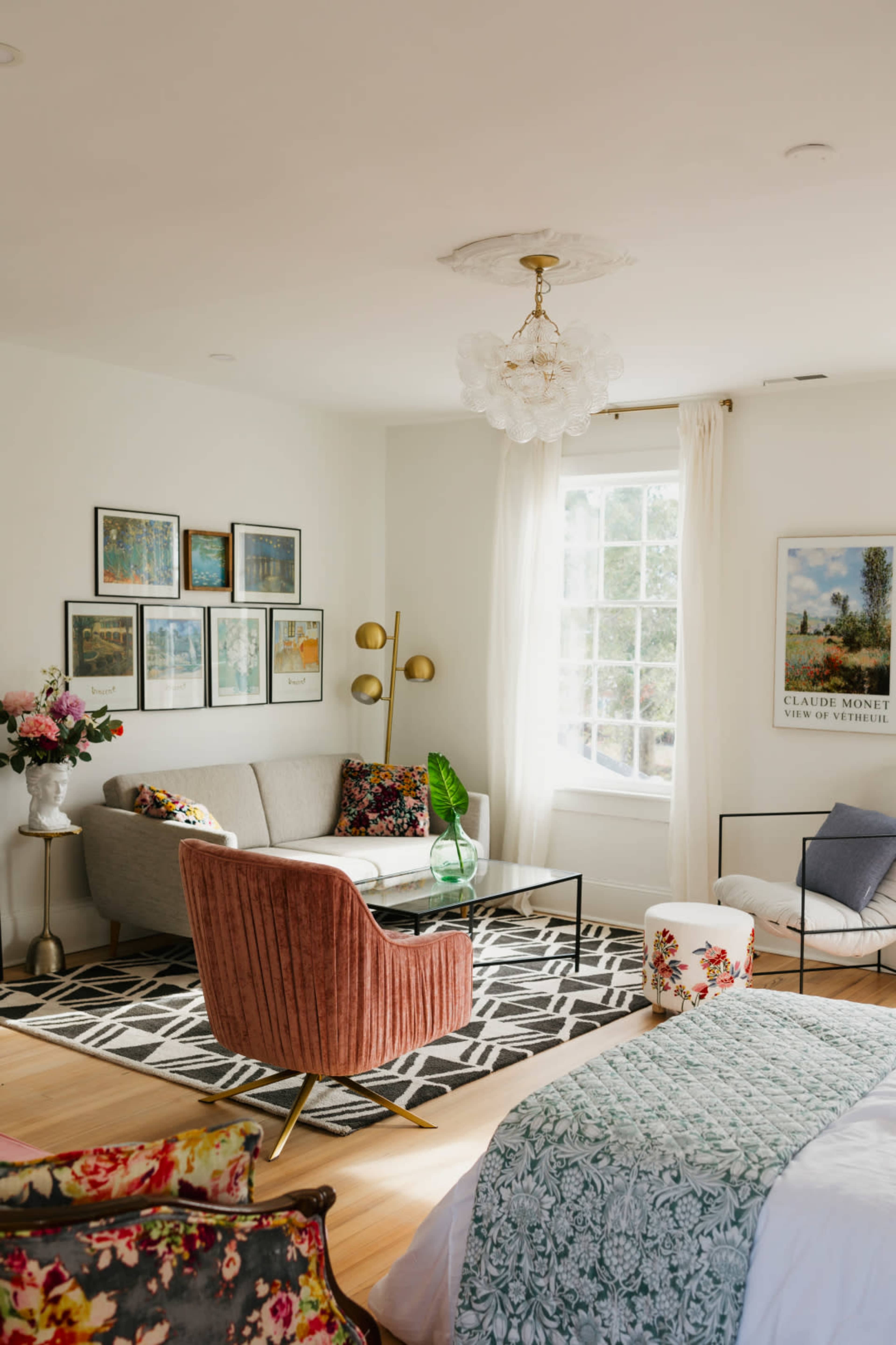 The image shows a bright, airy living room featuring a gray sofa, a pink armchair, a glass coffee table, and several framed artworks on the walls.