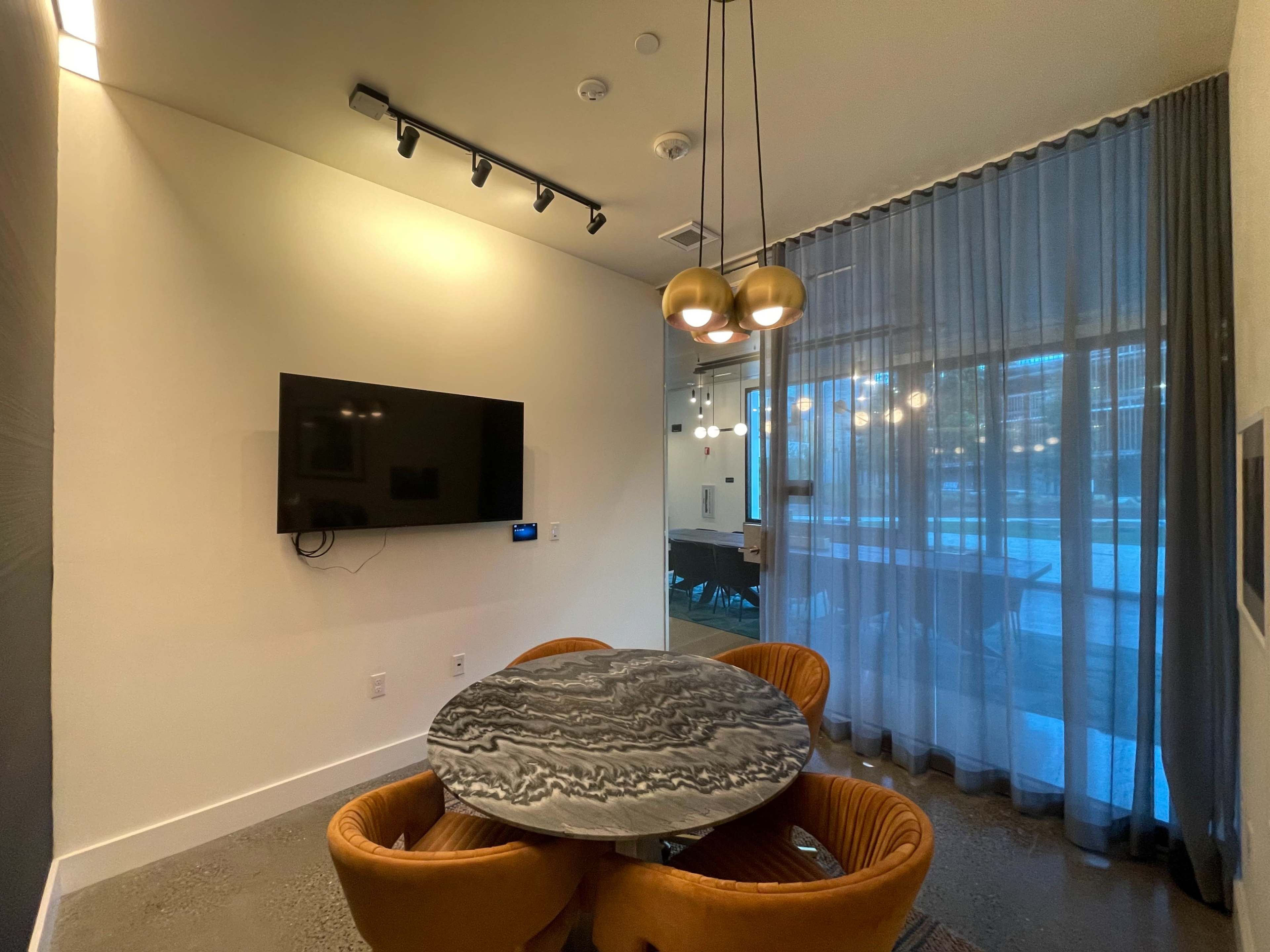 The scene features a modern room with a round marble table surrounded by three orange chairs, a wall-mounted television, and large windows adorned with sheer curtains.