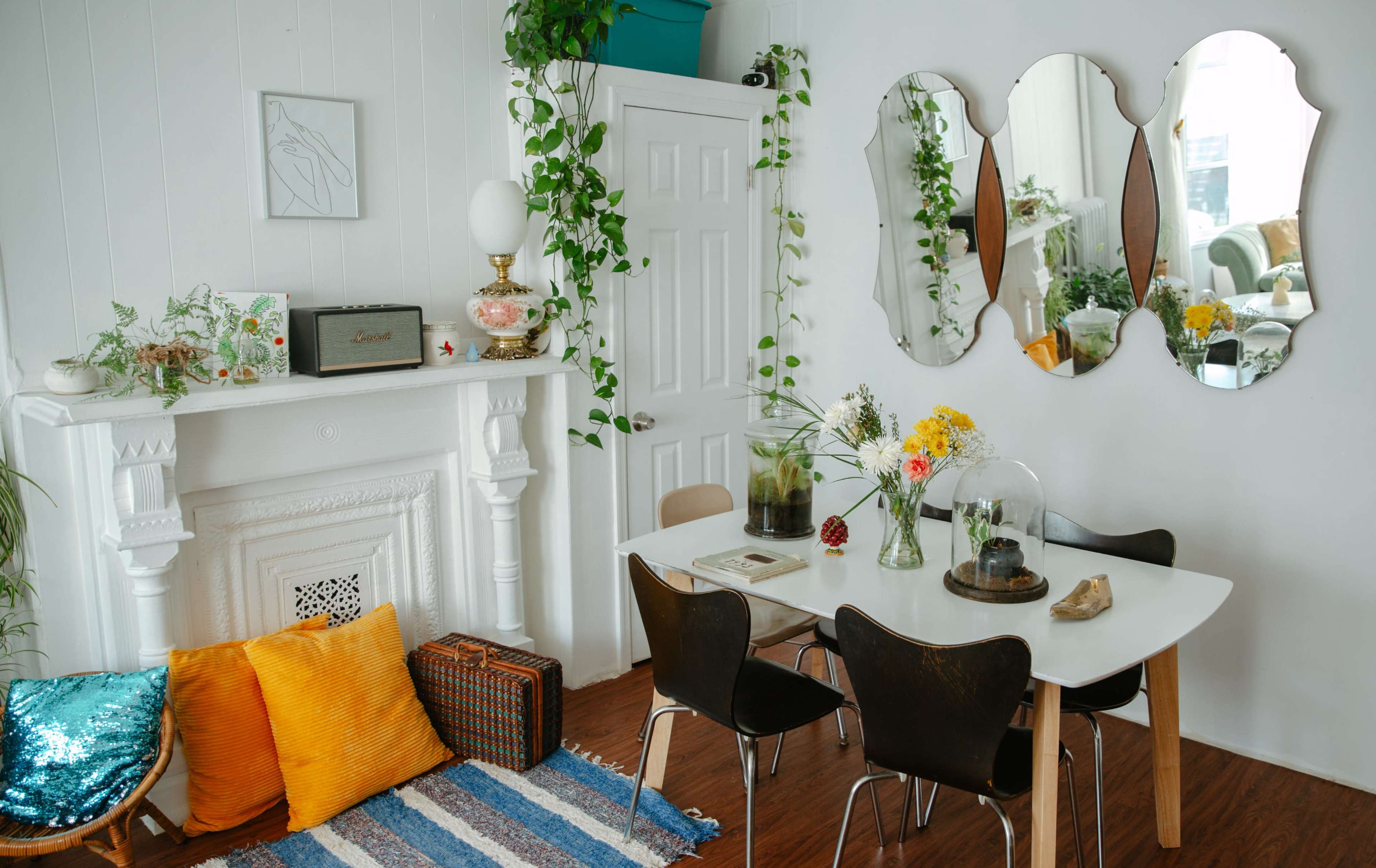 A bright dining area with a white table surrounded by black chairs, a decorative fireplace, potted plants, and three mirrors on the wall.