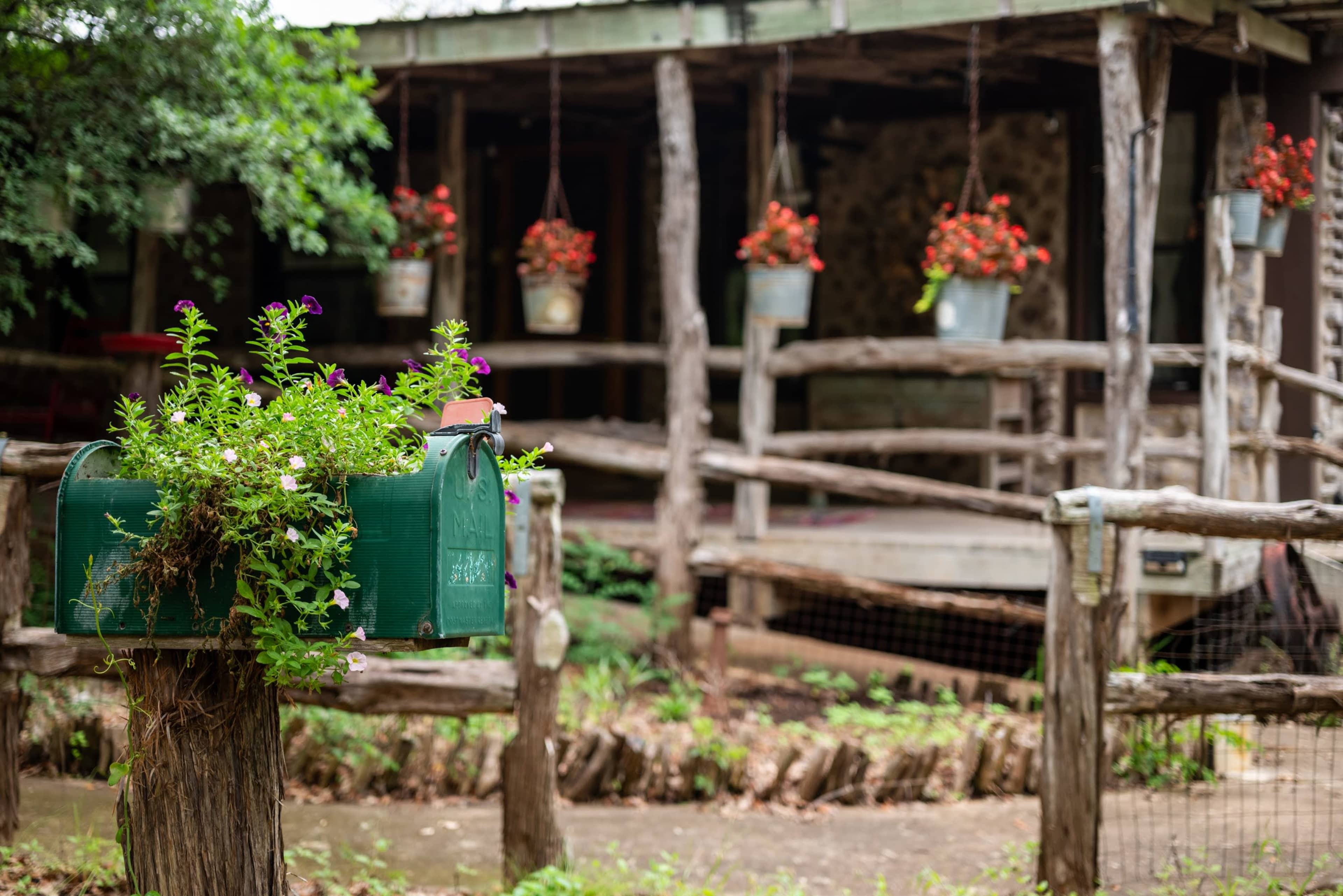 A green mailbox, overflowing with flowers, stands in the foreground of a rustic wooden porch adorned with hanging flower pots.