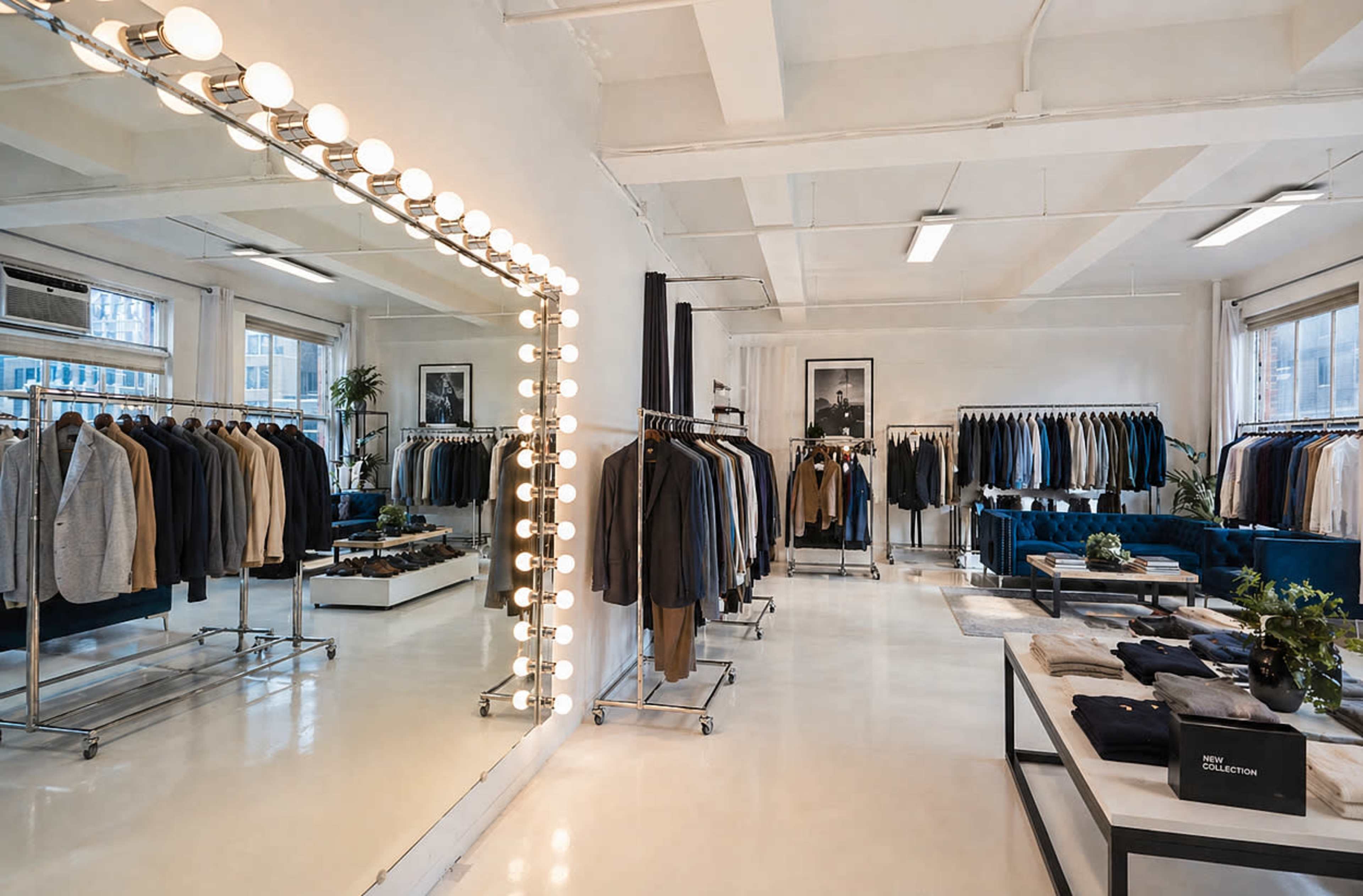 The image shows a modern clothing store with racks of suits, a large mirror with light bulbs, and a small seating area with blue furniture.