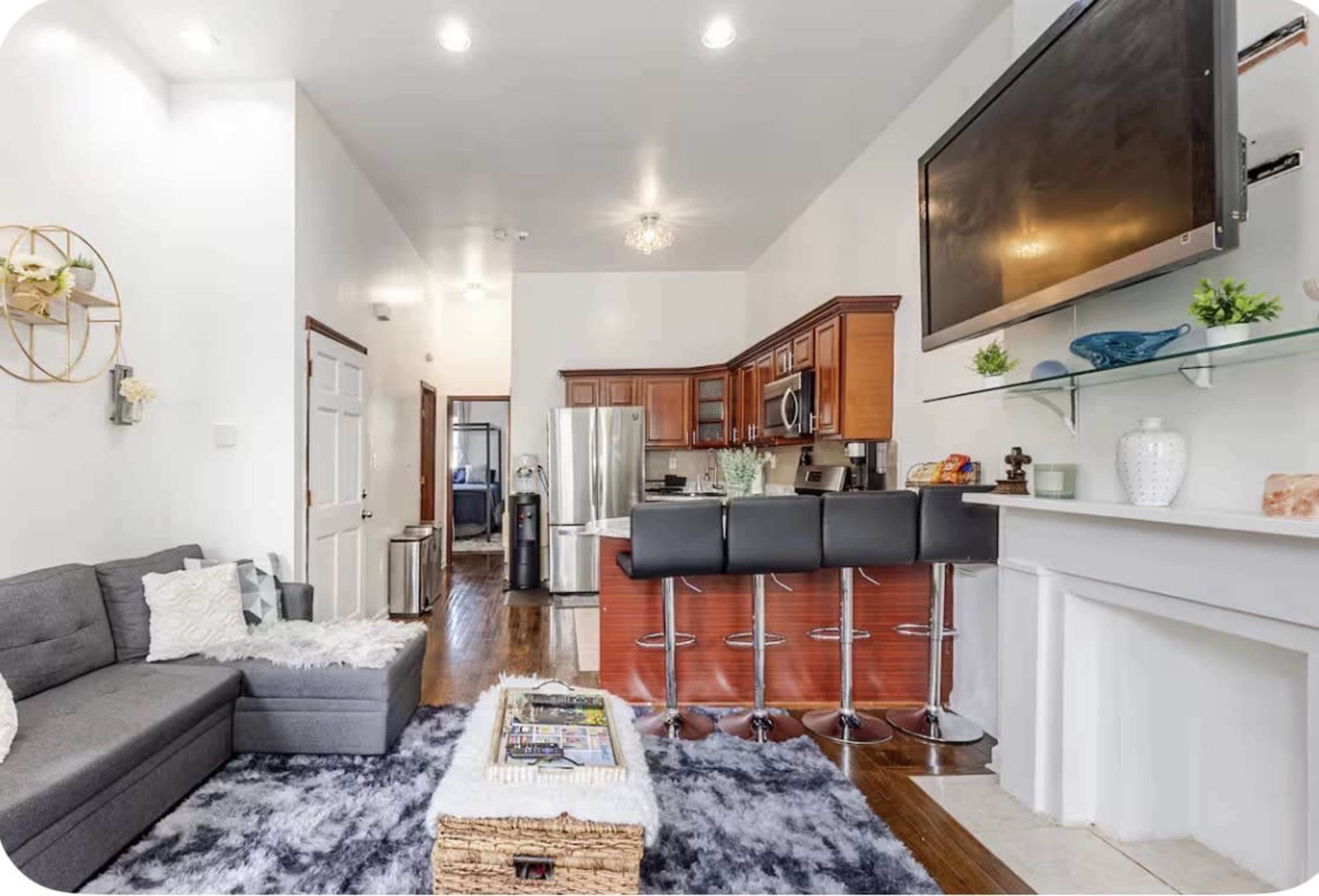 The image shows a modern living space featuring a gray sectional sofa, a kitchen with wooden cabinets, and a bar area with four black stools.