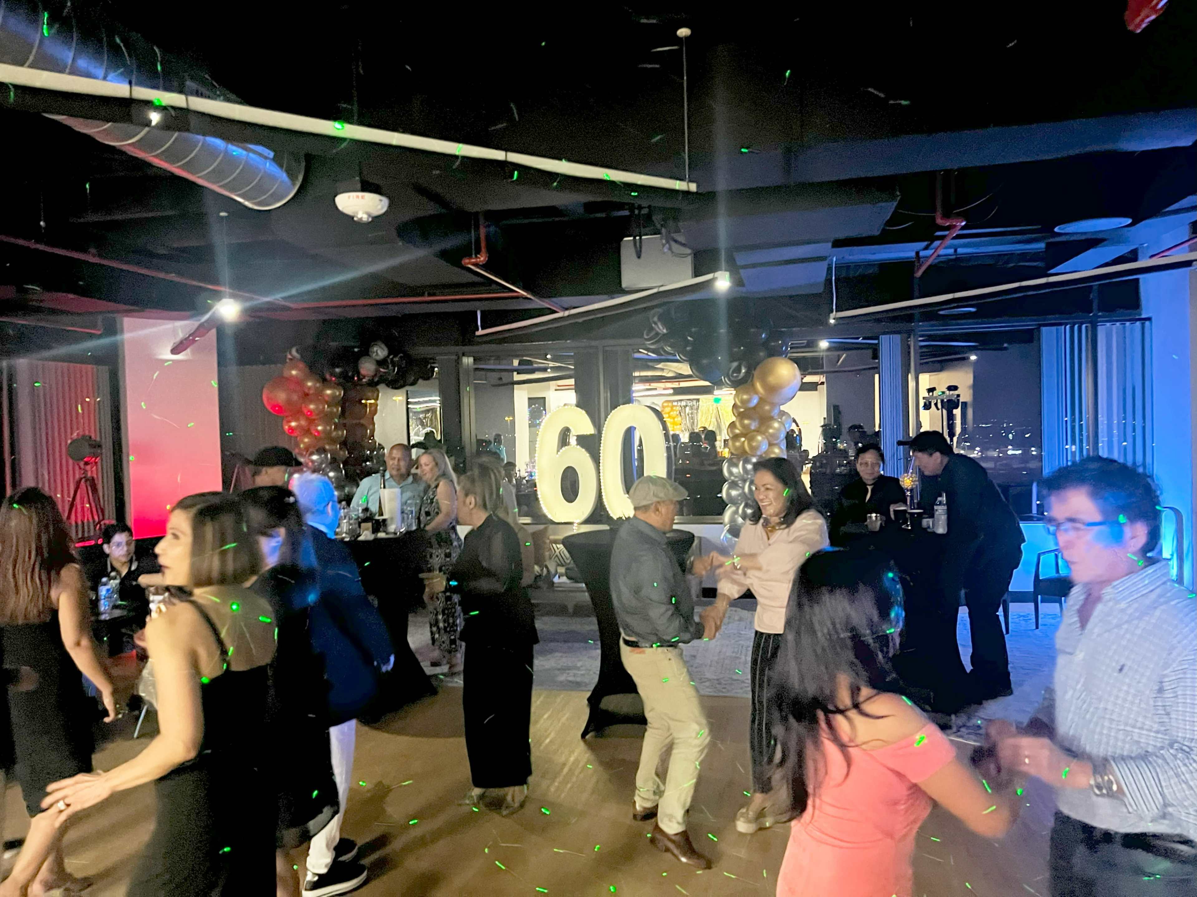 A lively party is taking place in an office space, featuring a large "60" centerpiece and guests dancing amid colorful lights.