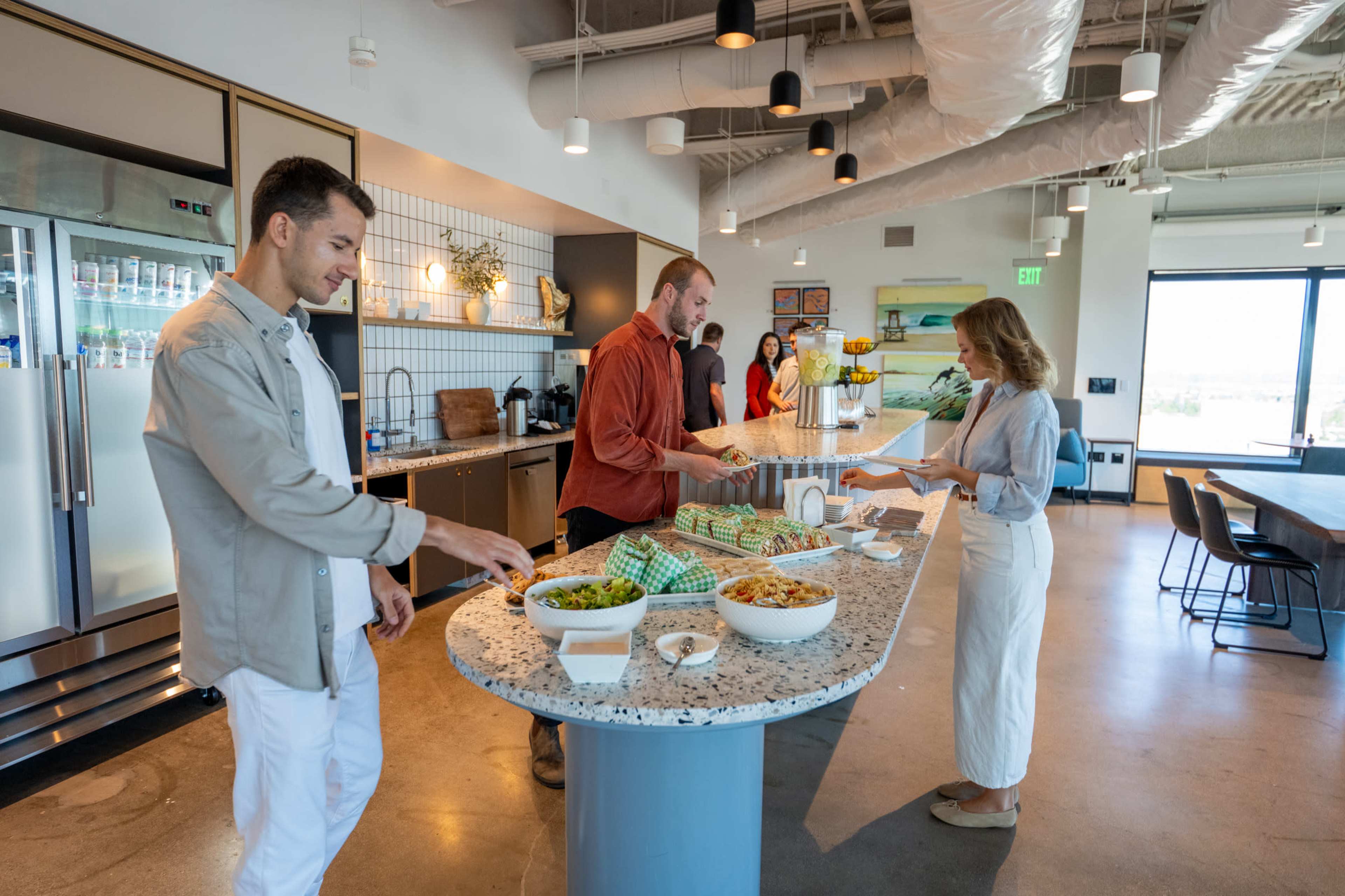 A group of four people is gathered around a large kitchen island, enjoying a spread of various dishes in a modern office space.