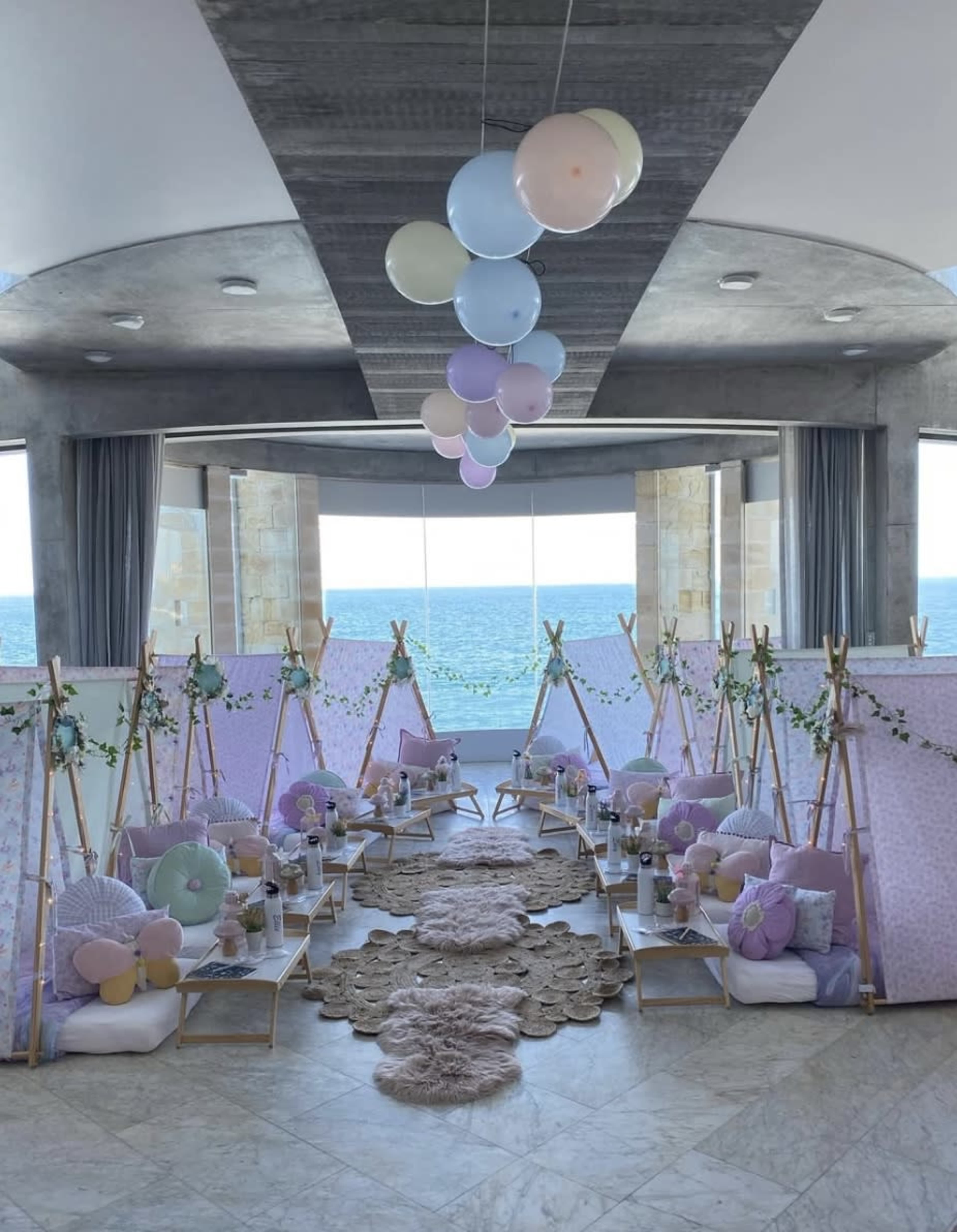 The scene features a brightly lit room with multiple pastel-colored tents arranged in a line, decorated with cushions and balloons, set against a backdrop of large windows overlooking the sea.