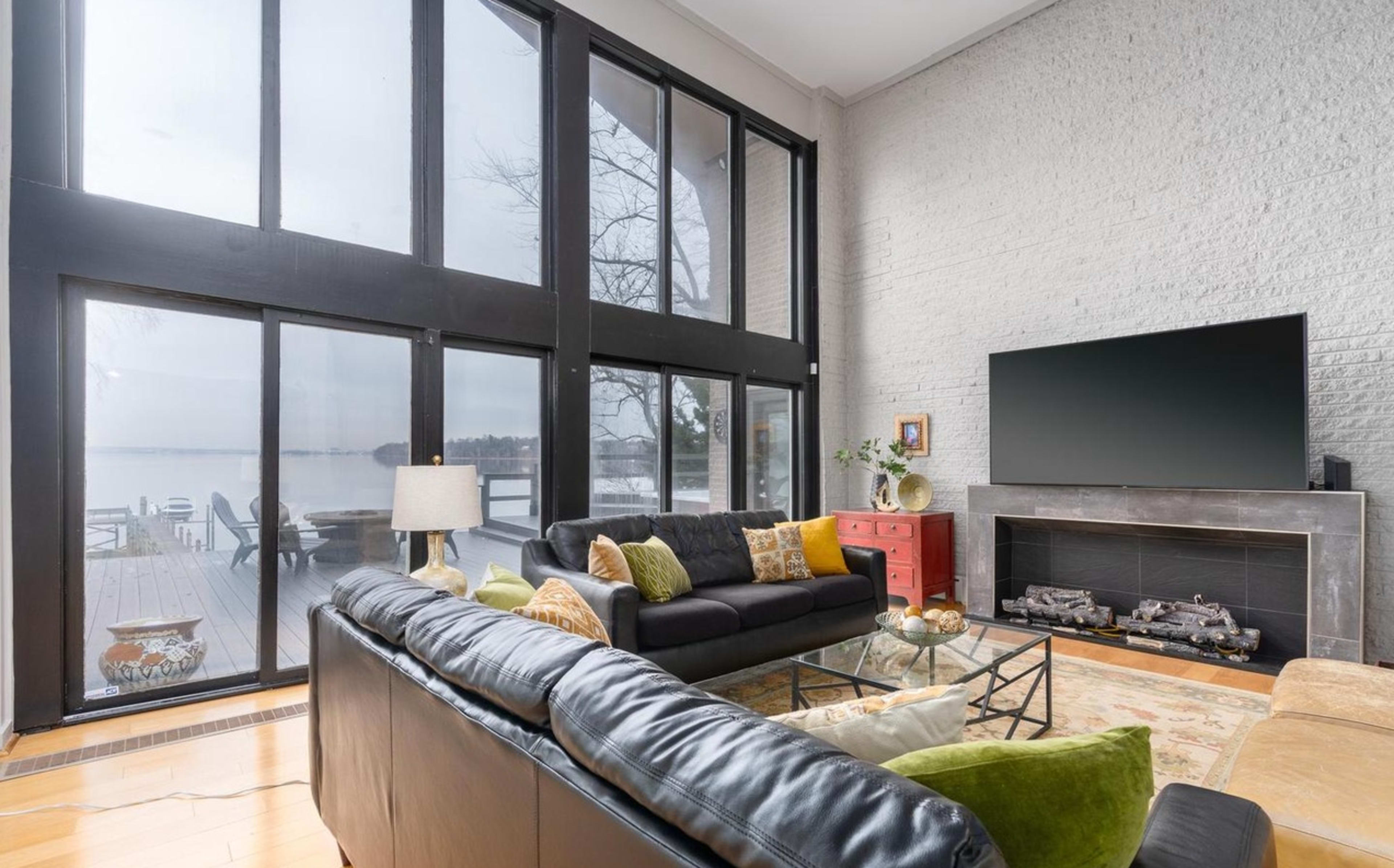 The image shows a modern living room with large windows overlooking a body of water, featuring a black leather sectional sofa, a glass coffee table, and a mounted television above a fireplace.
