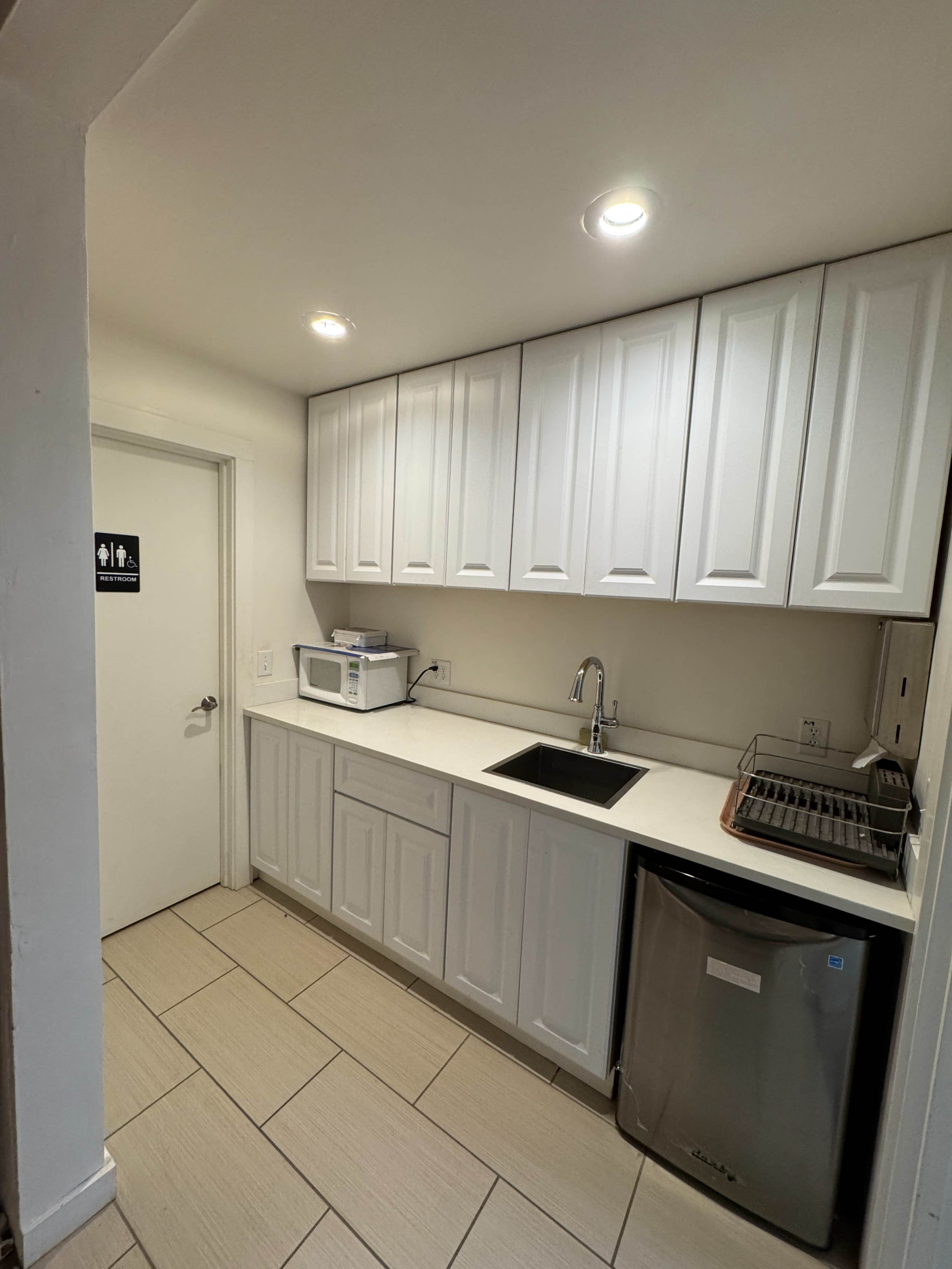 The image shows a small kitchen area with white cabinets, a sink, a microwave, and a small refrigerator.