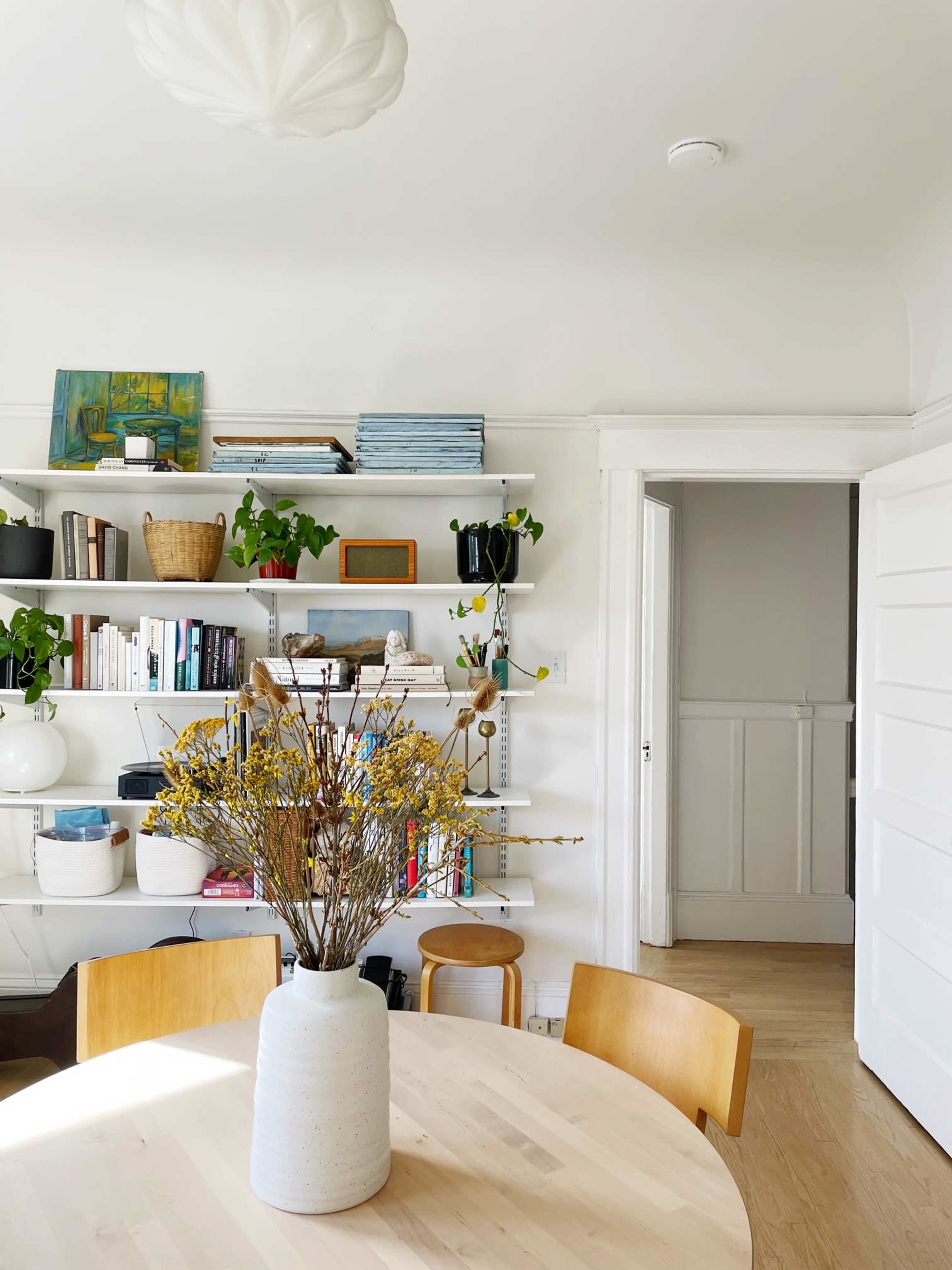A neutral-colored dining area features a round table with a vase of flowers, surrounded by wooden chairs, and bookshelves filled with plants and various items against a white wall.