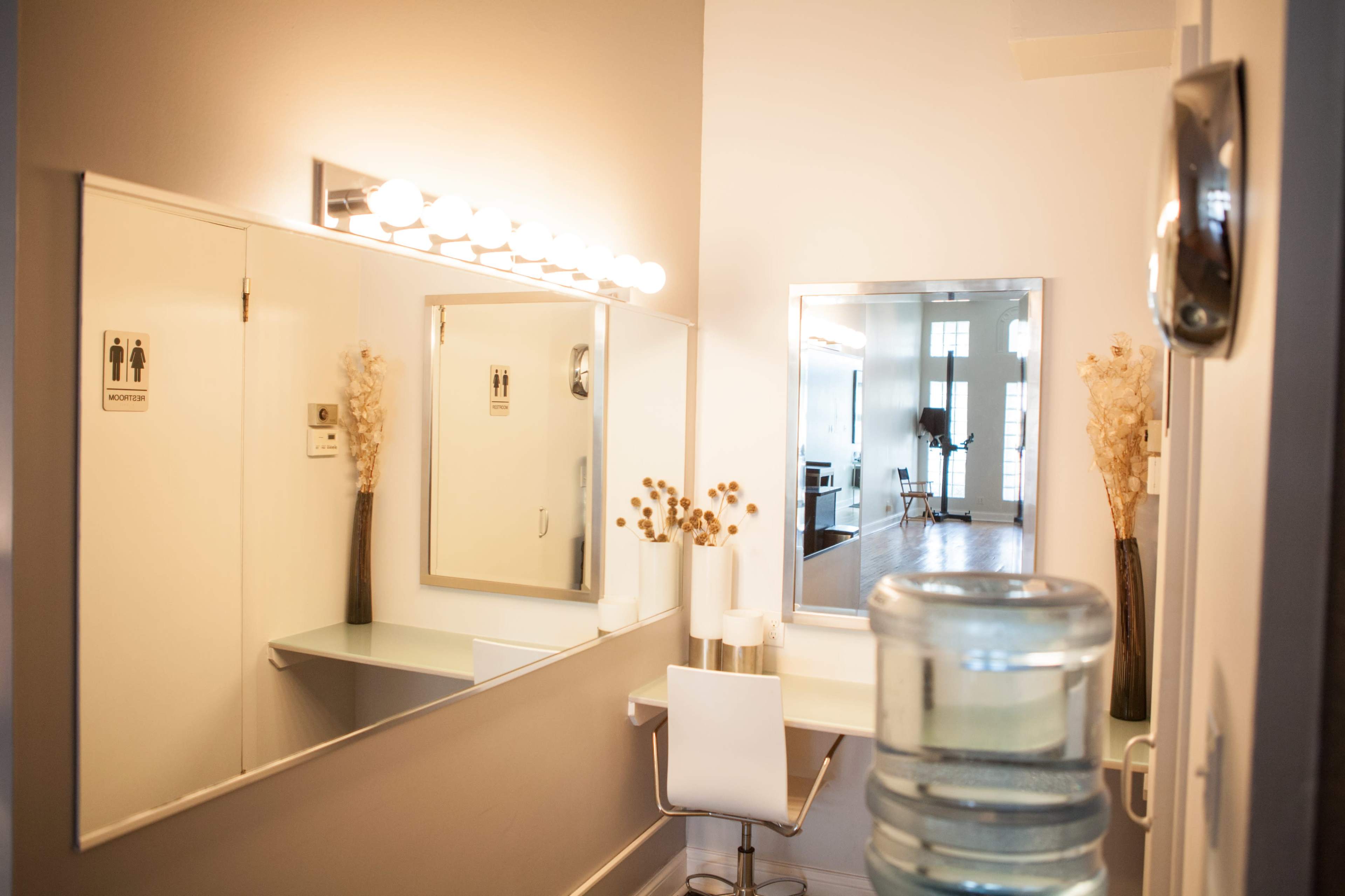 The image shows a well-lit vanity area with a mirror, a chair at a table, and decorative plants, adjacent to a door marked with restroom signs.