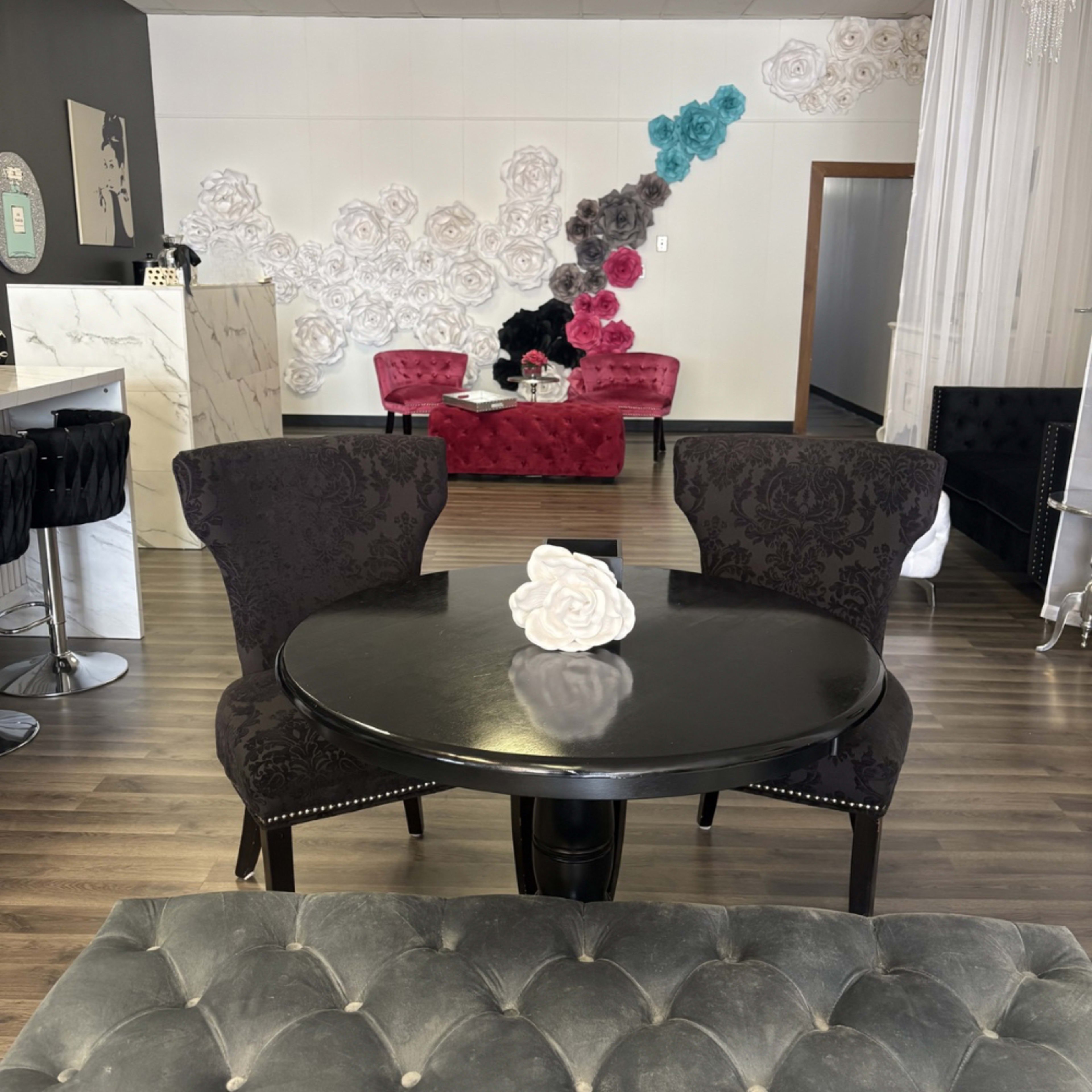 A black circular table with two ornate chairs sits in the center of a room decorated with white and colored paper flowers on the wall, alongside plush red seating.