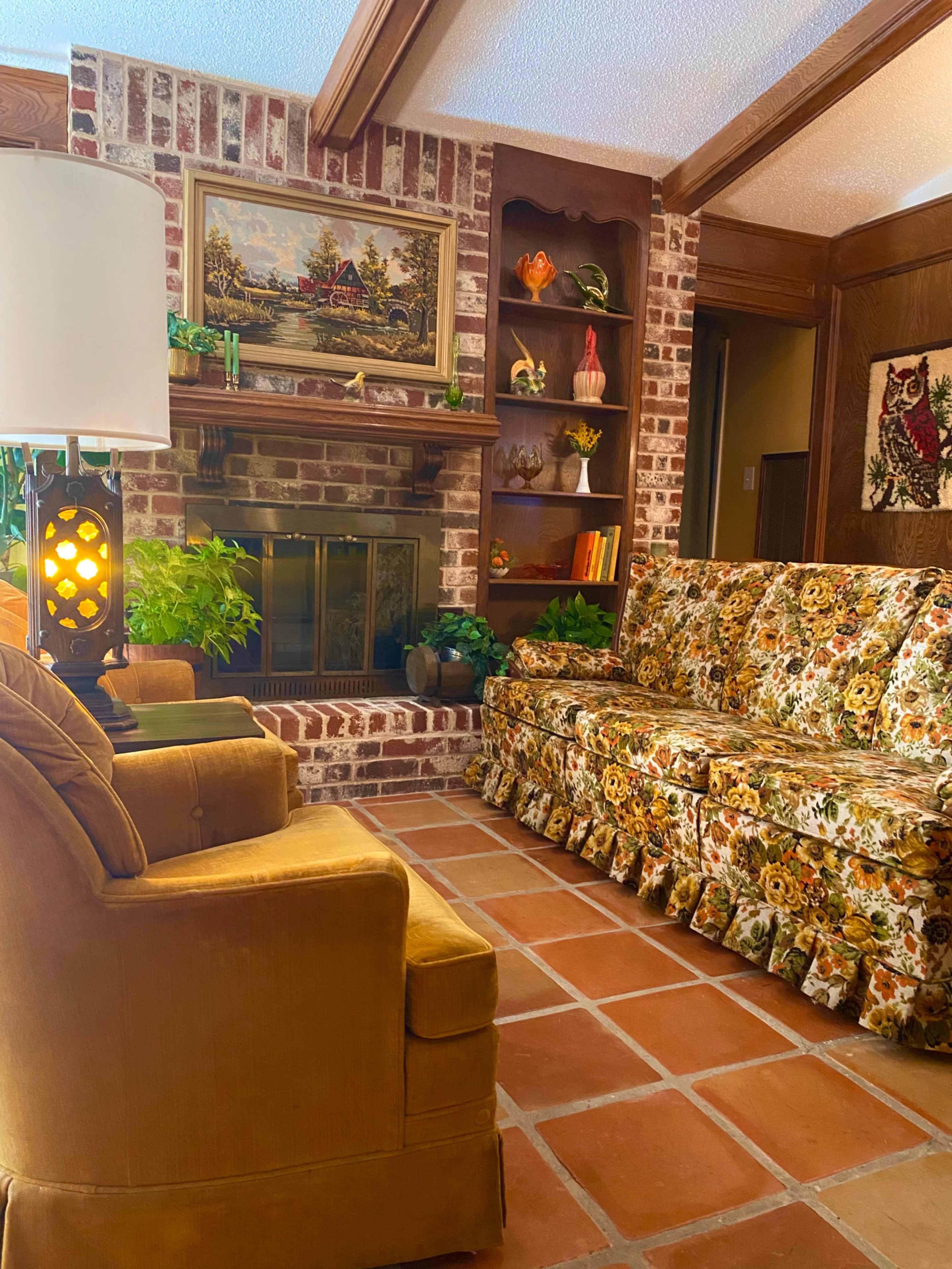 The image shows a living room with a brick fireplace, floral-patterned sofa, and a lamp beside an armchair.