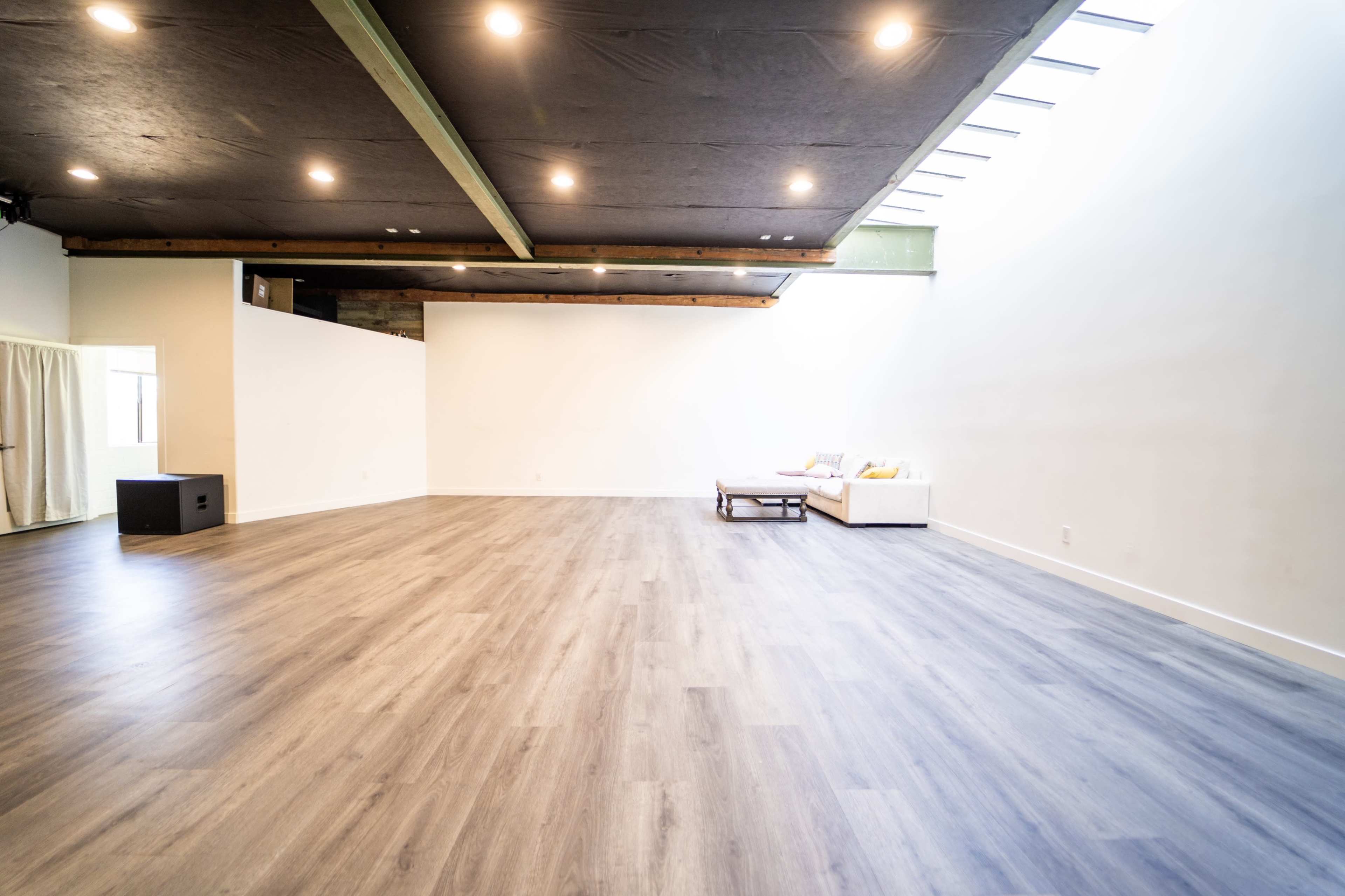 The image shows a spacious, empty room with wooden flooring and large windows allowing natural light to enter.