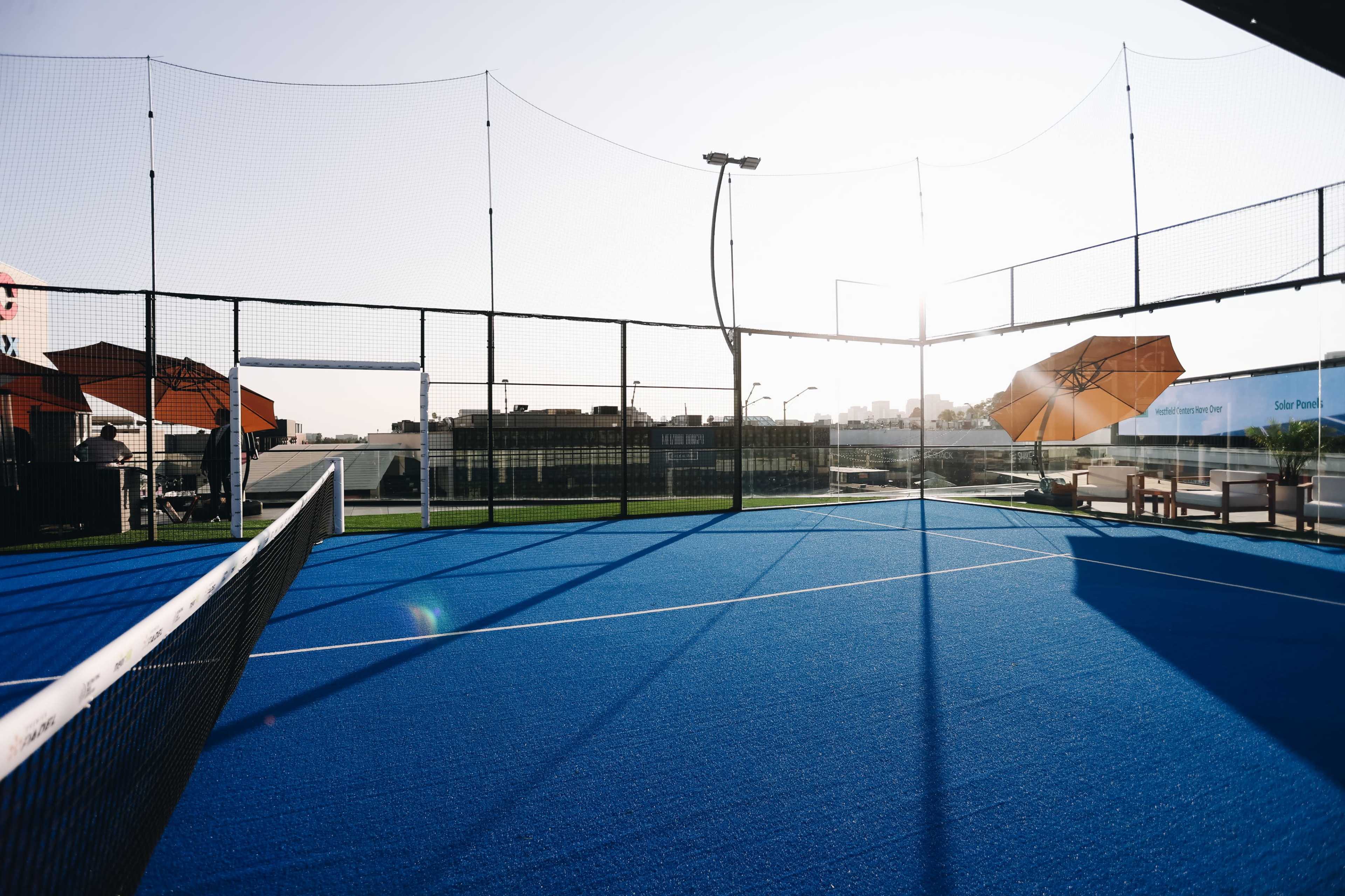 The image shows a blue tennis court with a net, surrounded by fences and two umbrellas in the background, under a bright sky.