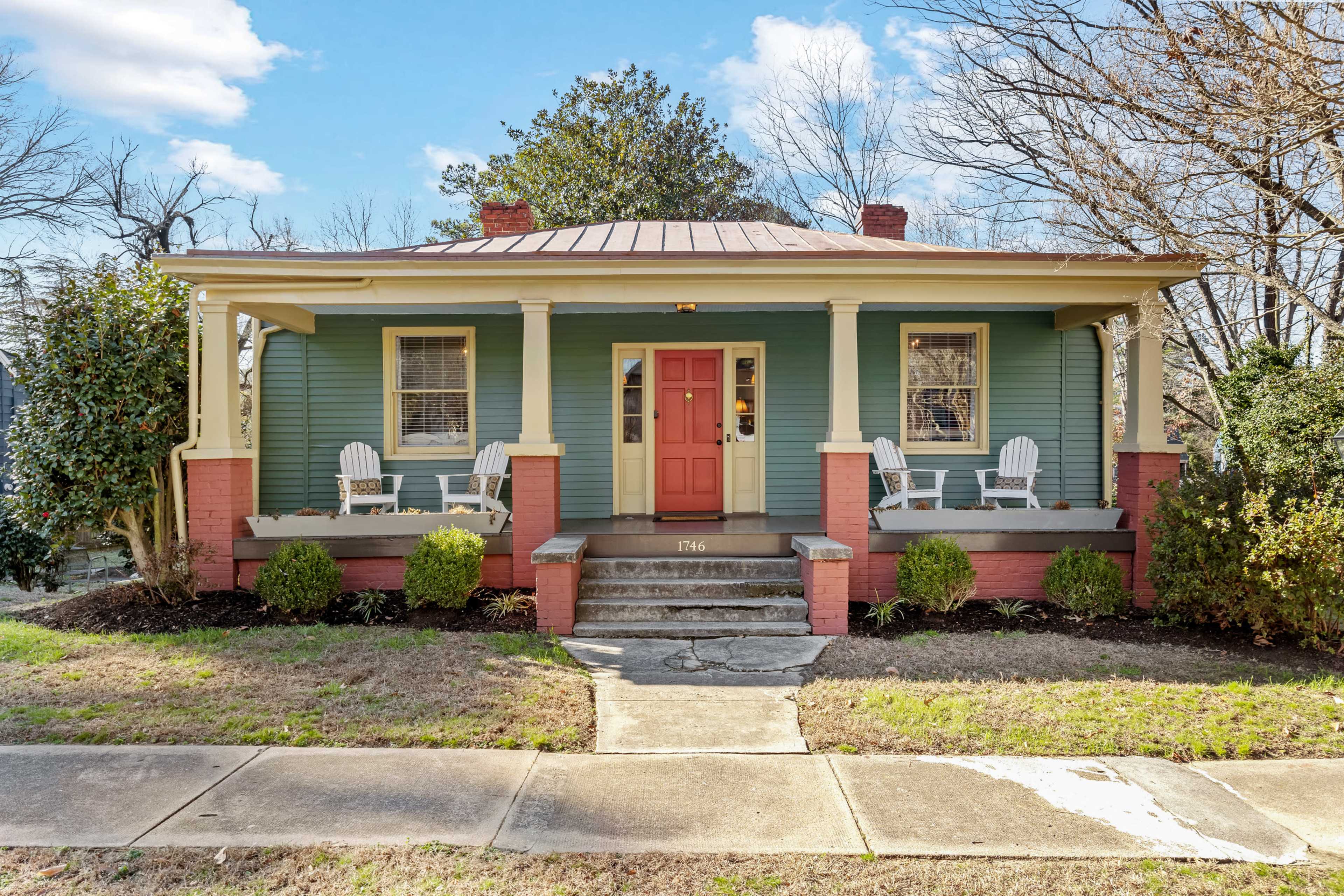 A green house with a red door and white rocking chairs on the porch, surrounded by well-maintained landscaping.