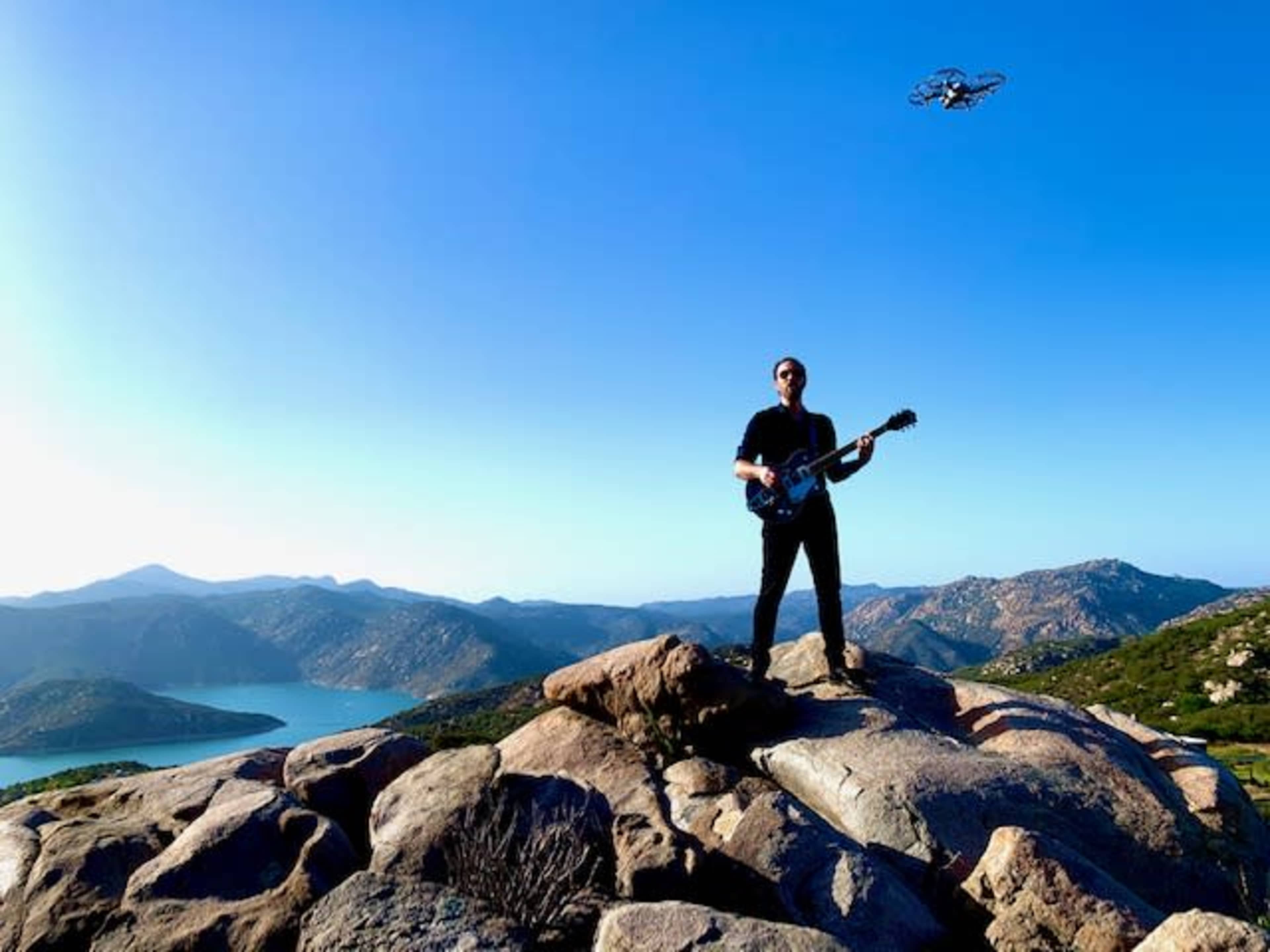A musician playing an electric guitar stands on rocky terrain with a scenic view of a lake and hills, while a drone hovers above.