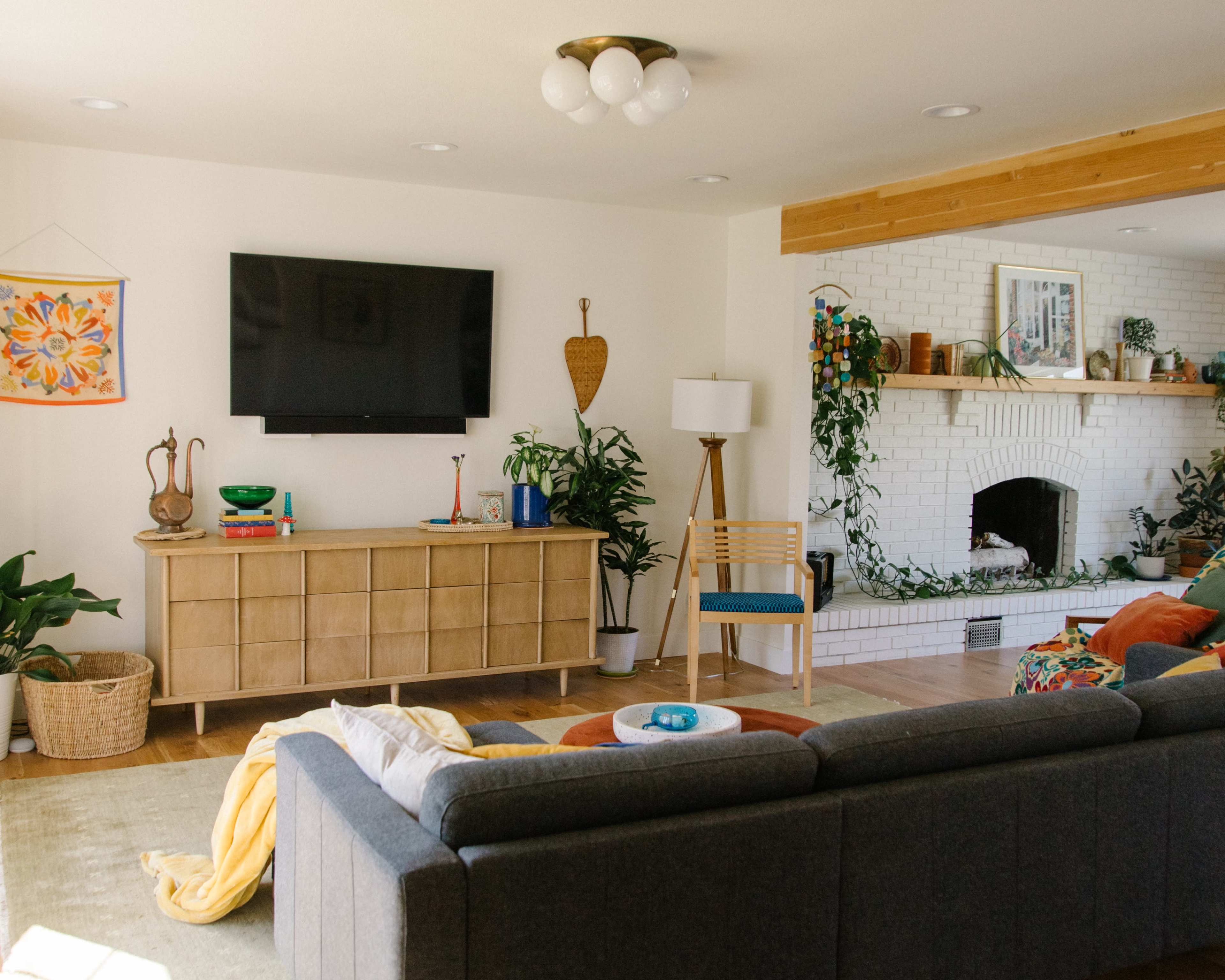 A modern living room with a television mounted on the wall, a wooden sideboard, a plant-filled fireplace, and a seating area that includes a gray couch and a wooden chair.