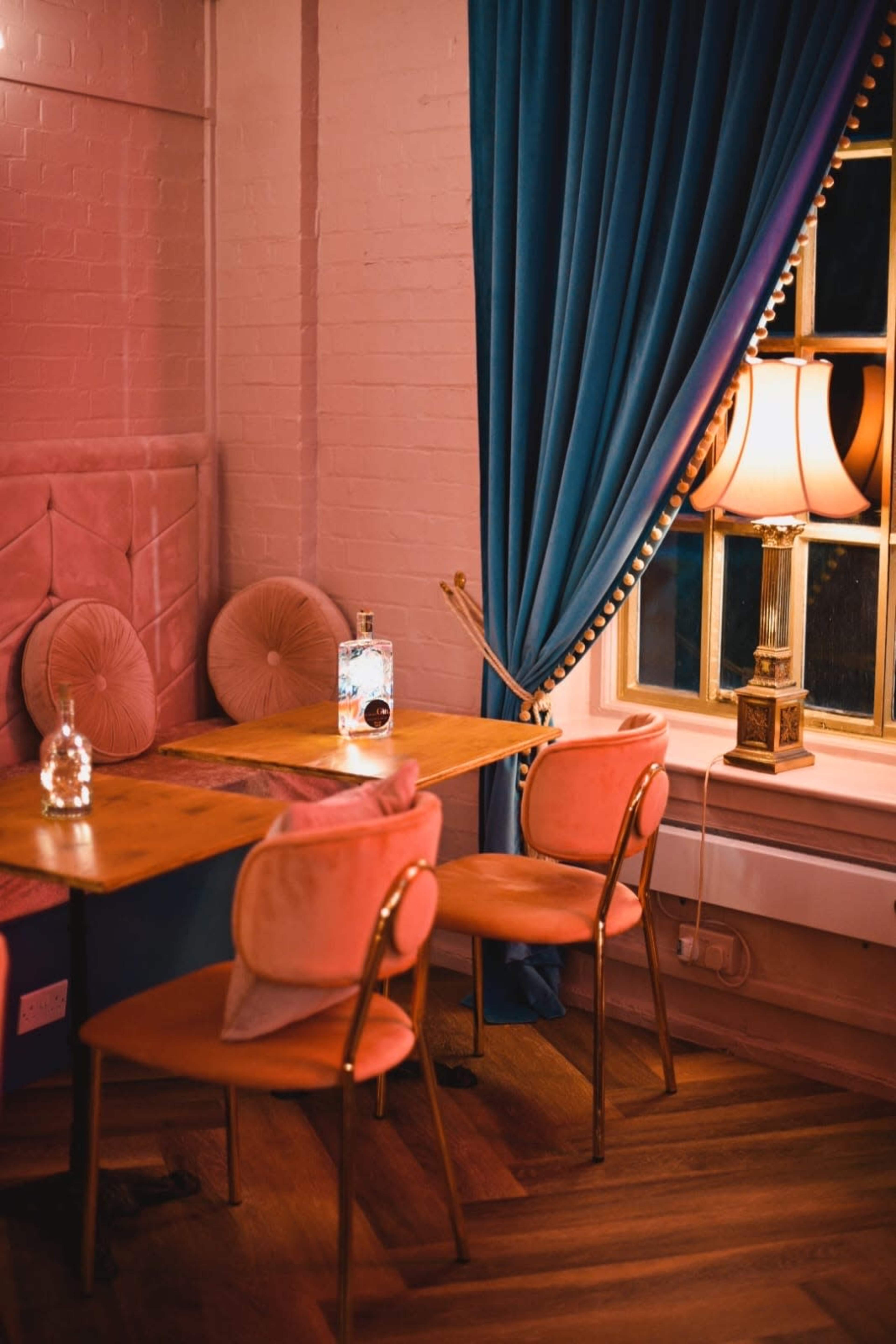 A table with a bottle of water and a glass sits beside plush, pink velvet chairs in a softly lit room featuring a blue curtain and a lamp.