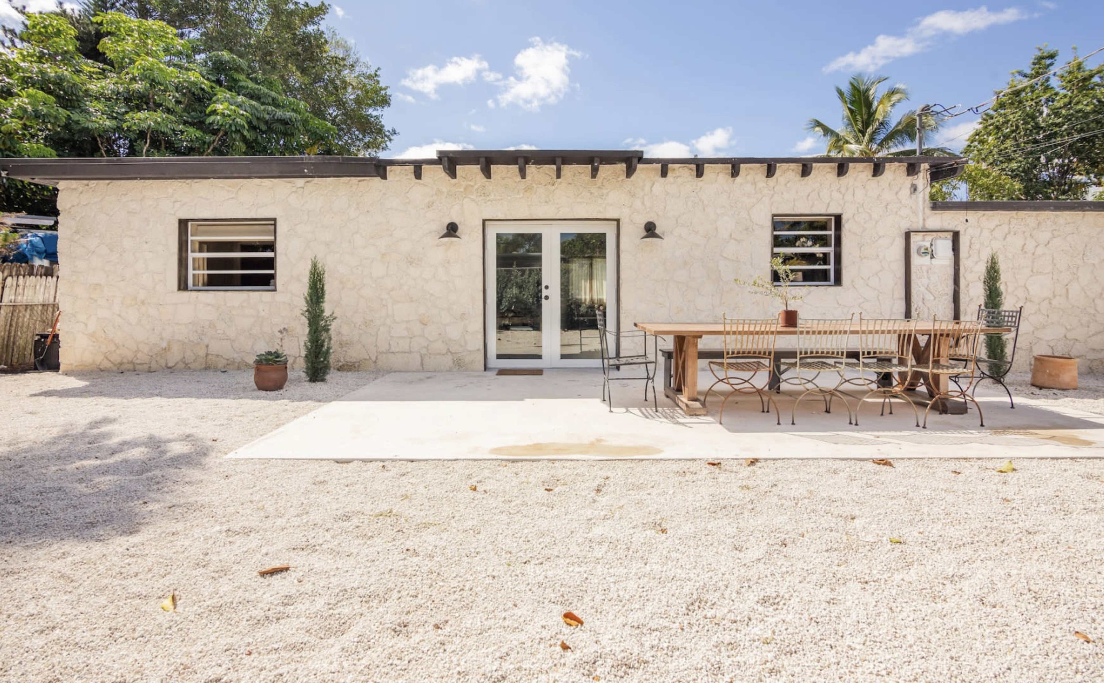 A stone-clad building features large glass doors opening to a patio with a long wooden dining table and chairs, surrounded by gravel and greenery.