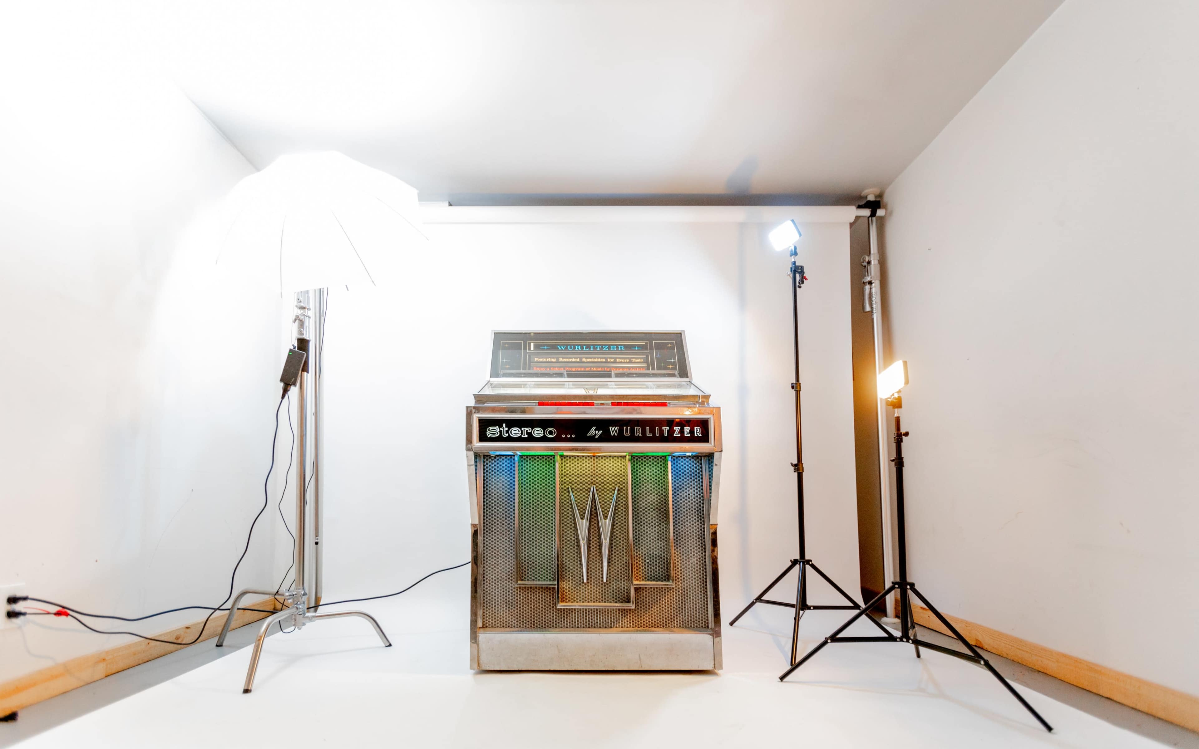 A vintage jukebox sits in a well-lit studio space, flanked by two light stands and an umbrella softbox.