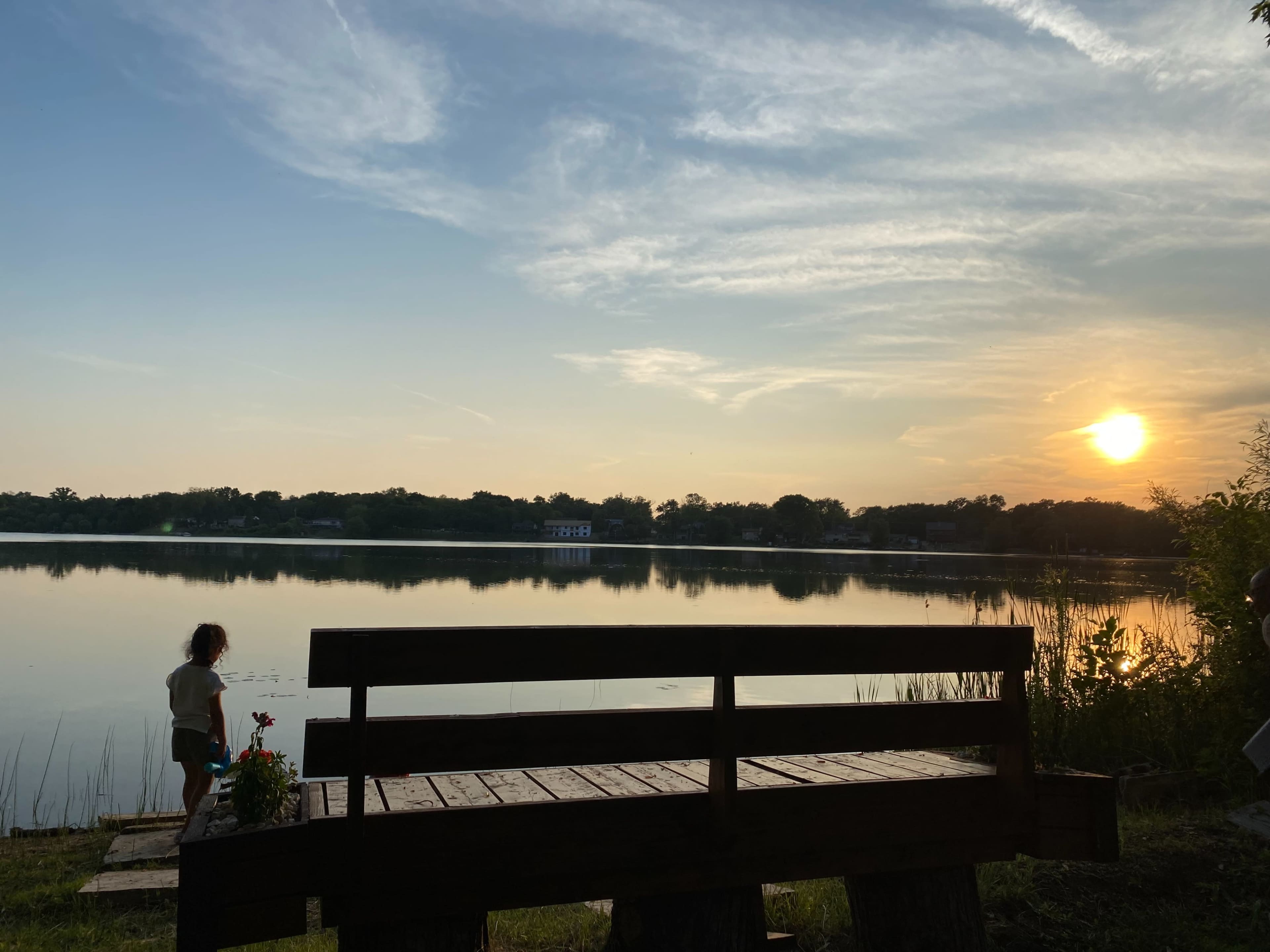 A child stands near the water's edge at sunset, with a bench in the foreground and a calm lake reflecting the light.