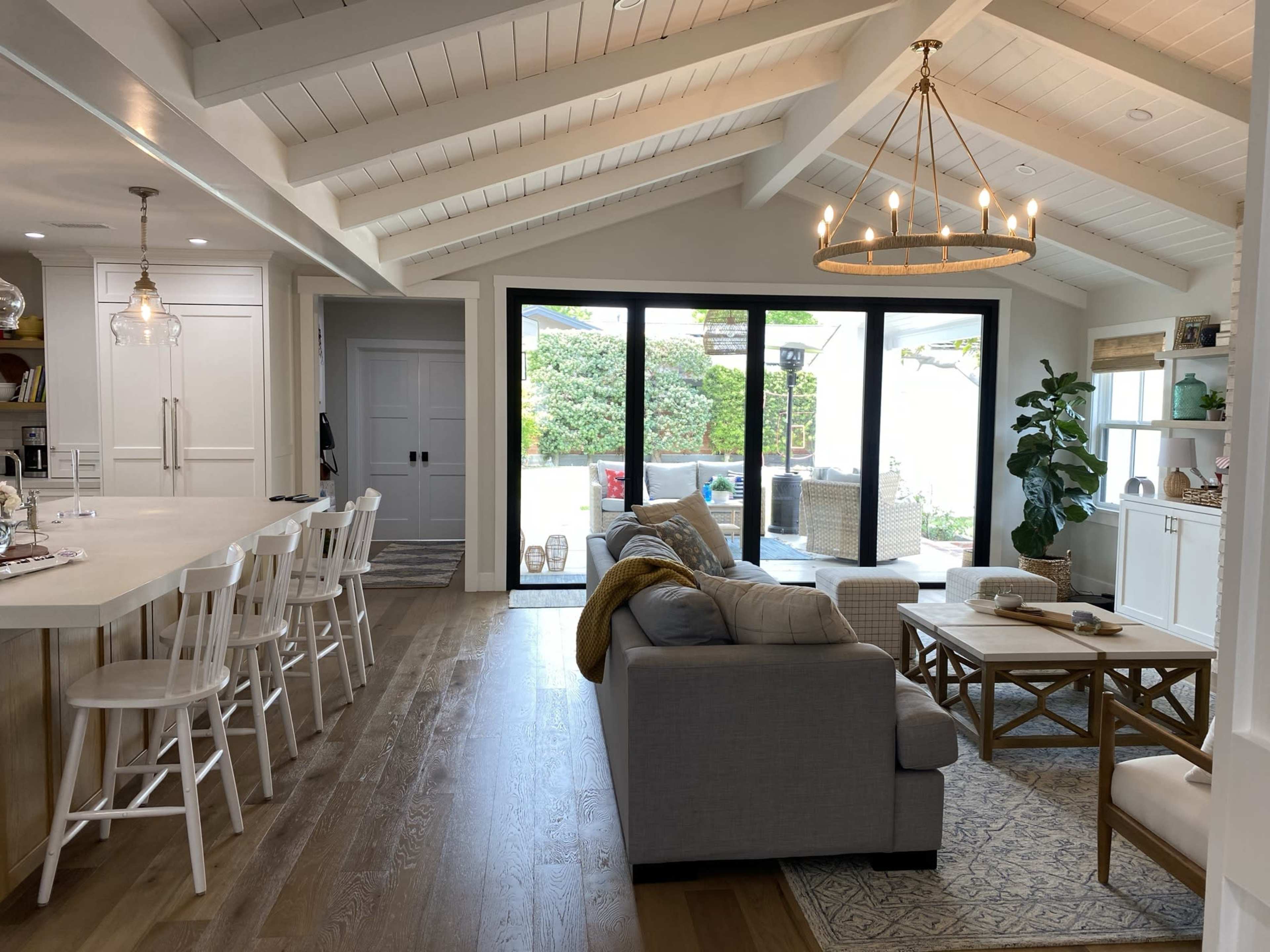 A modern living room with a connected kitchen featuring white cabinetry, a large central island with bar stools, and a spacious outdoor patio visible through sliding glass doors.