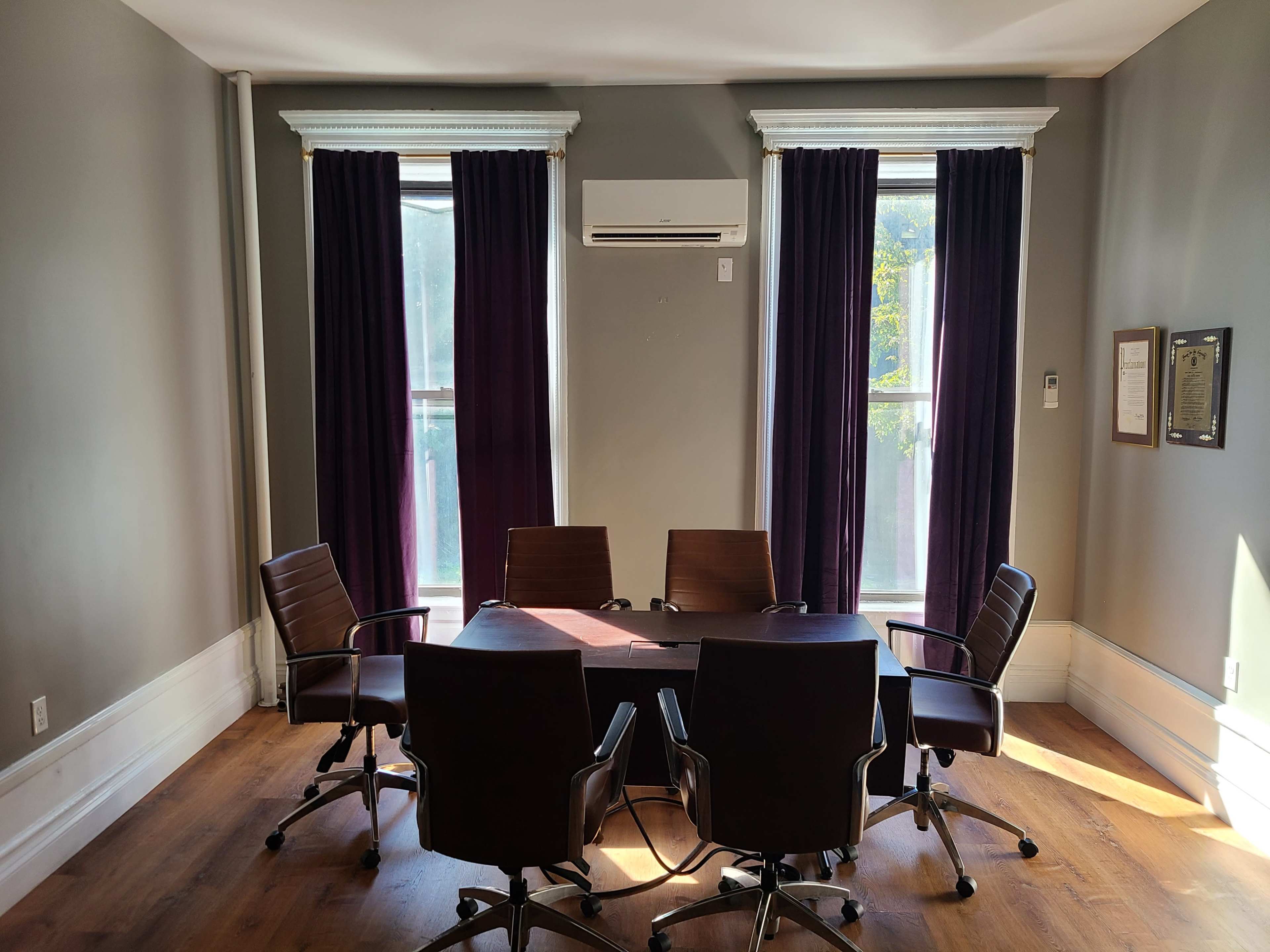 A conference room features a table surrounded by five chairs, with large windows dressed in dark curtains on either side.