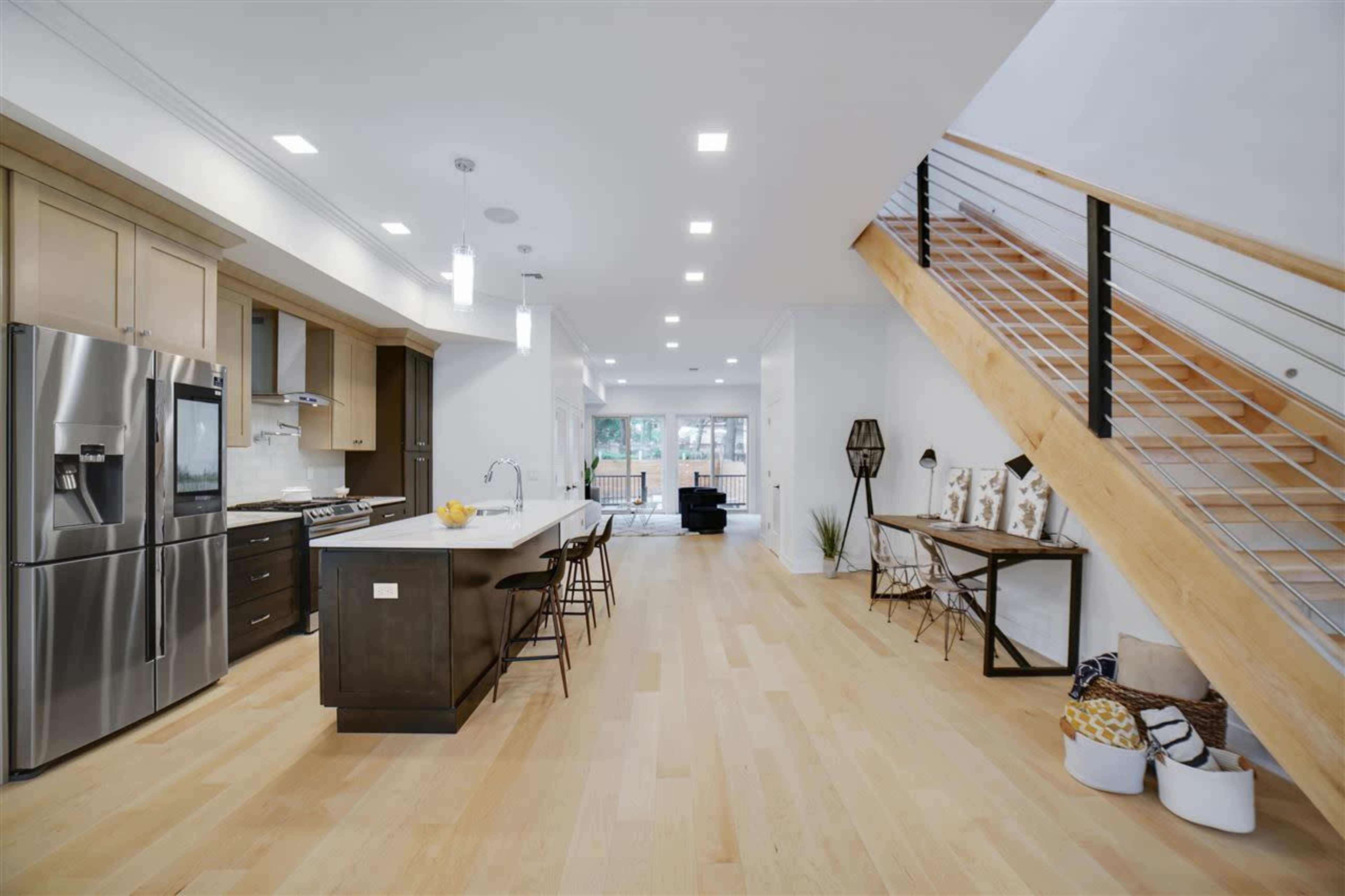 A modern kitchen with a kitchen island, stainless steel appliances, and a staircase leading to an upper level, with large windows allowing natural light.