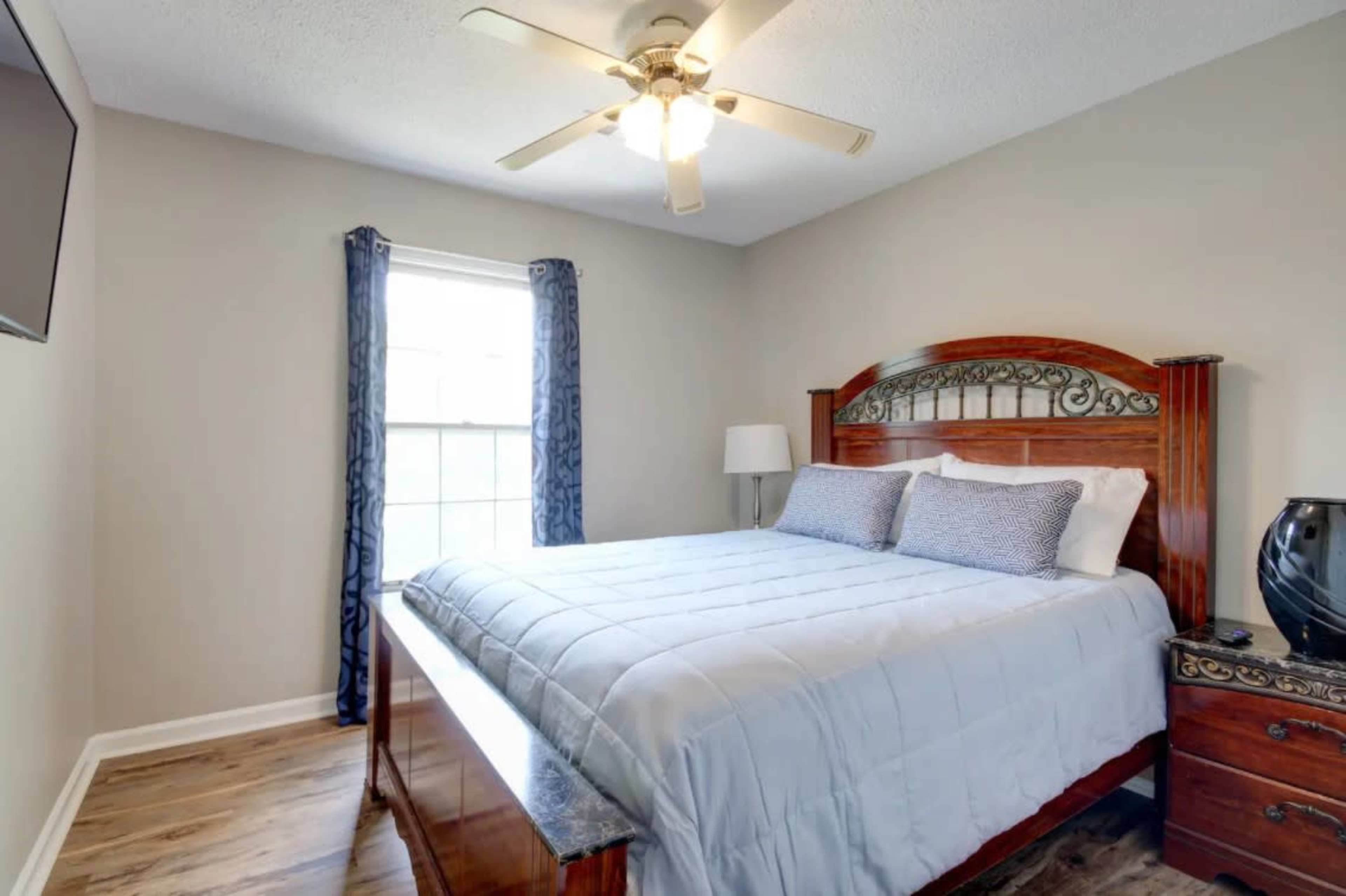 A well-lit bedroom features a wooden bed with a light blue quilt, a decorative lamp, and a window with drapes.