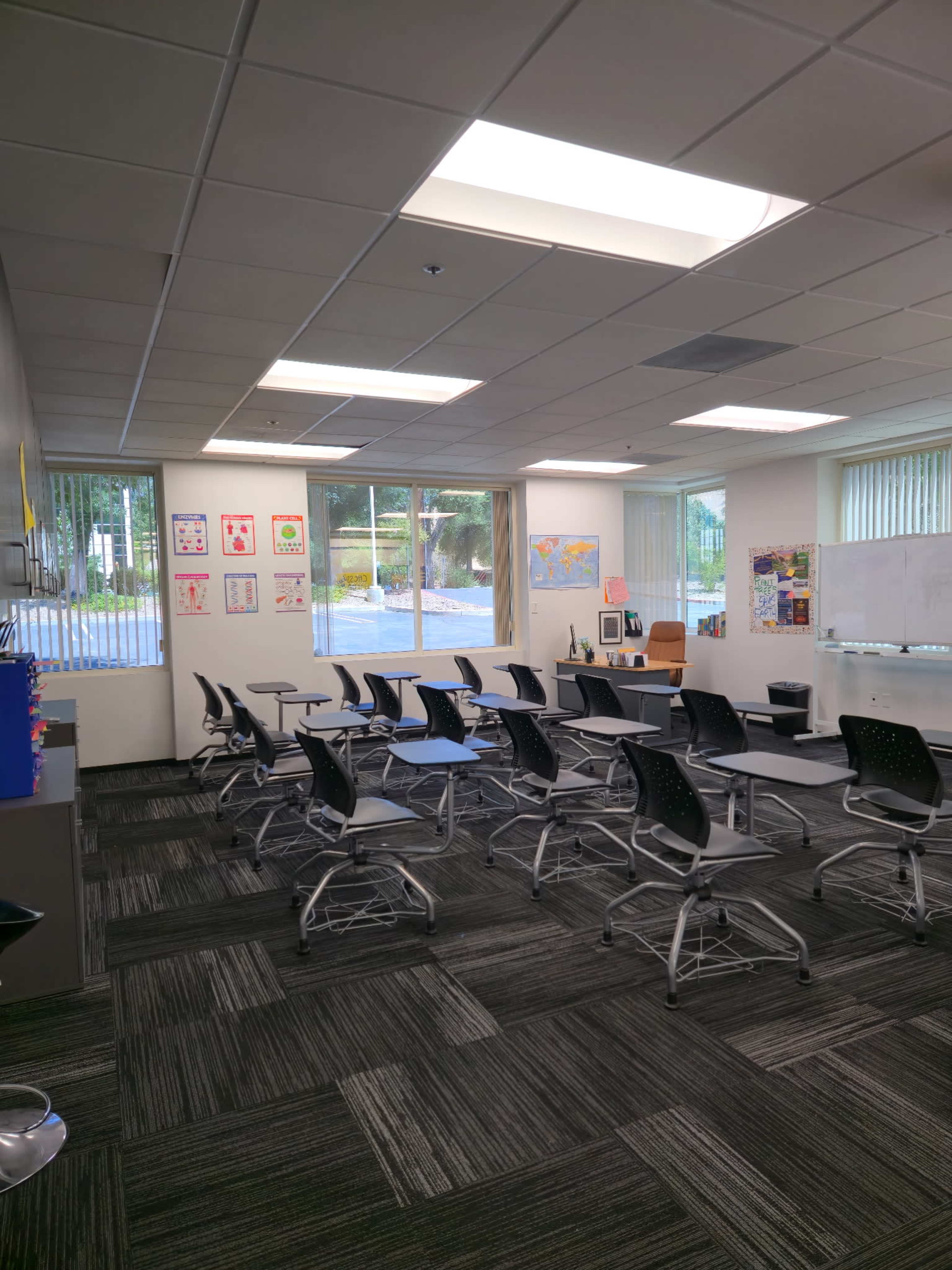 A classroom features rows of movable chairs and tables, large windows allowing natural light, and educational materials displayed on the walls.