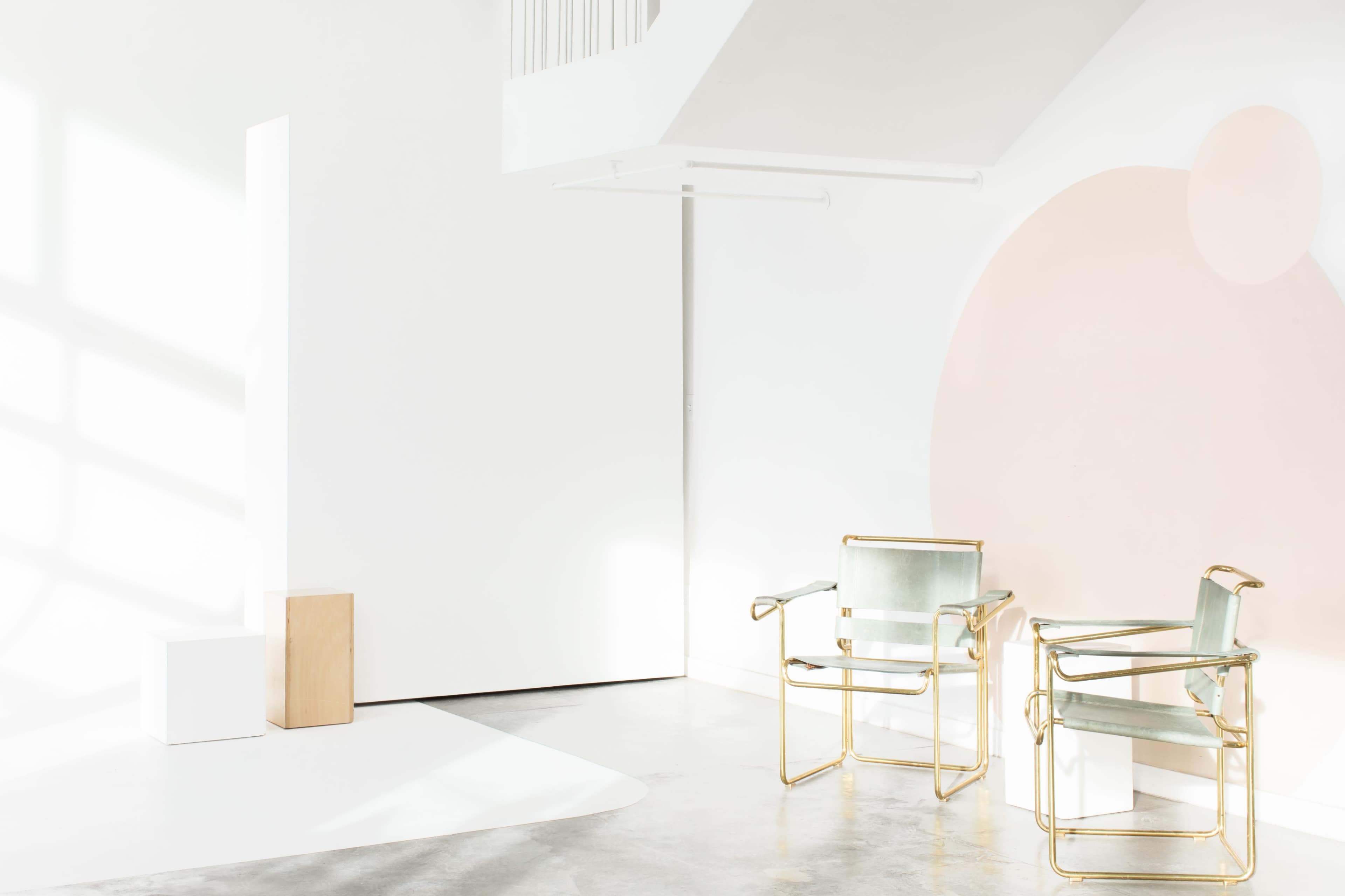 A minimalistic interior with two gold-framed chairs next to a large white wall, accented by a light pink circular design.