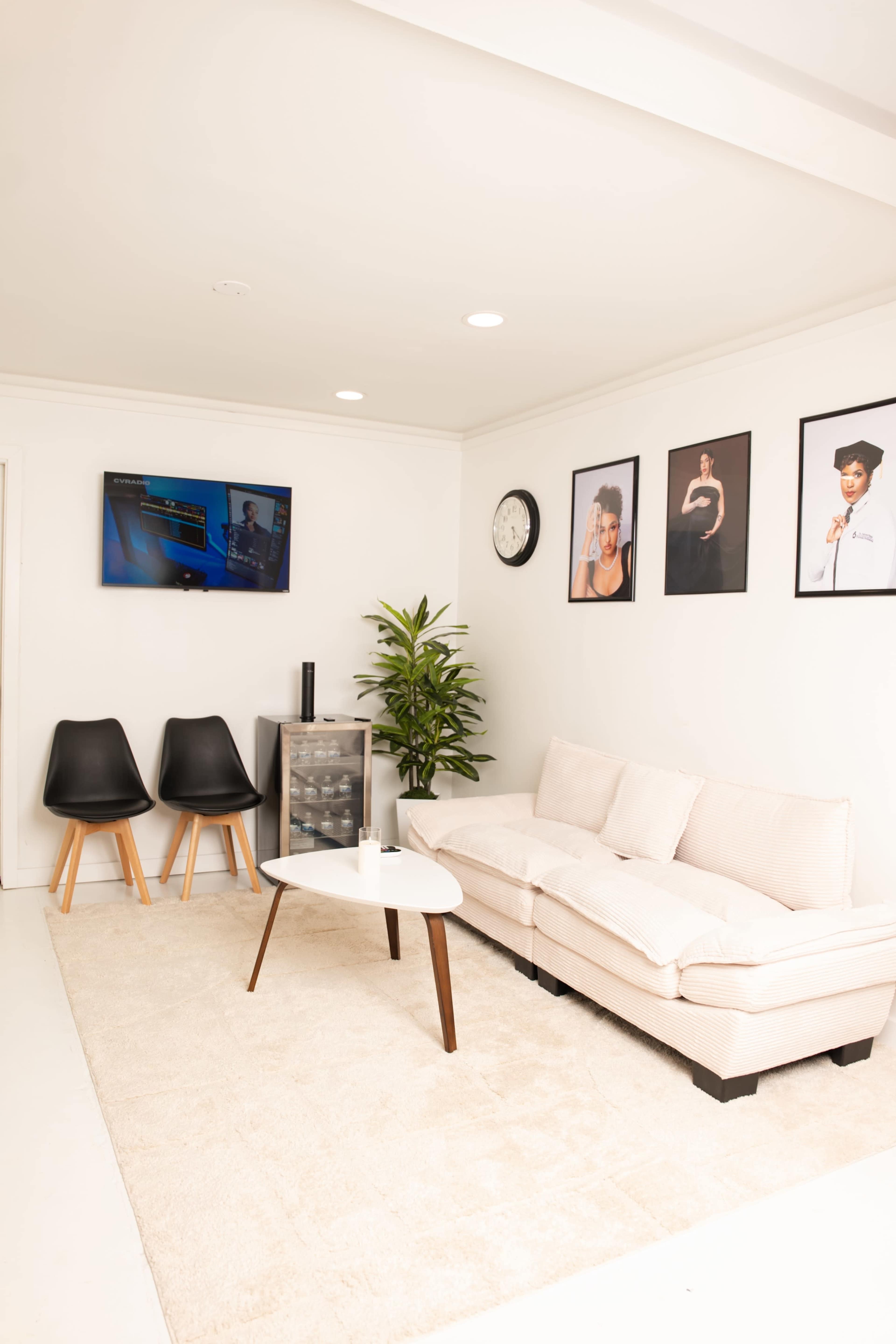 The image shows a modern, minimalist living area featuring a light-colored sofa, a coffee table, two black chairs, a plant, and framed pictures on the wall, along with a television mounted above a small cabinet.
