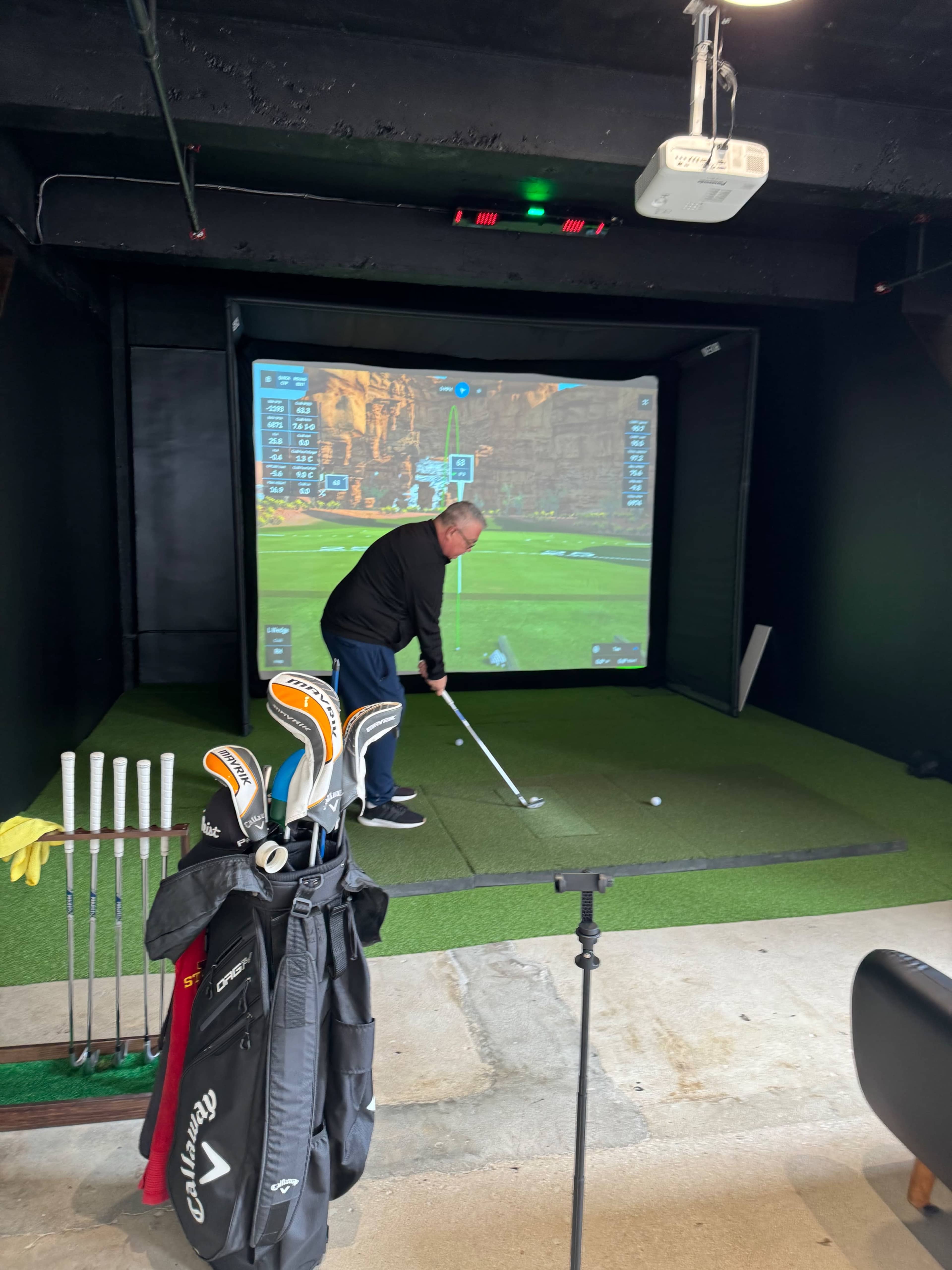 A person is taking a golf swing in a simulation room with a large screen displaying a virtual golf course.