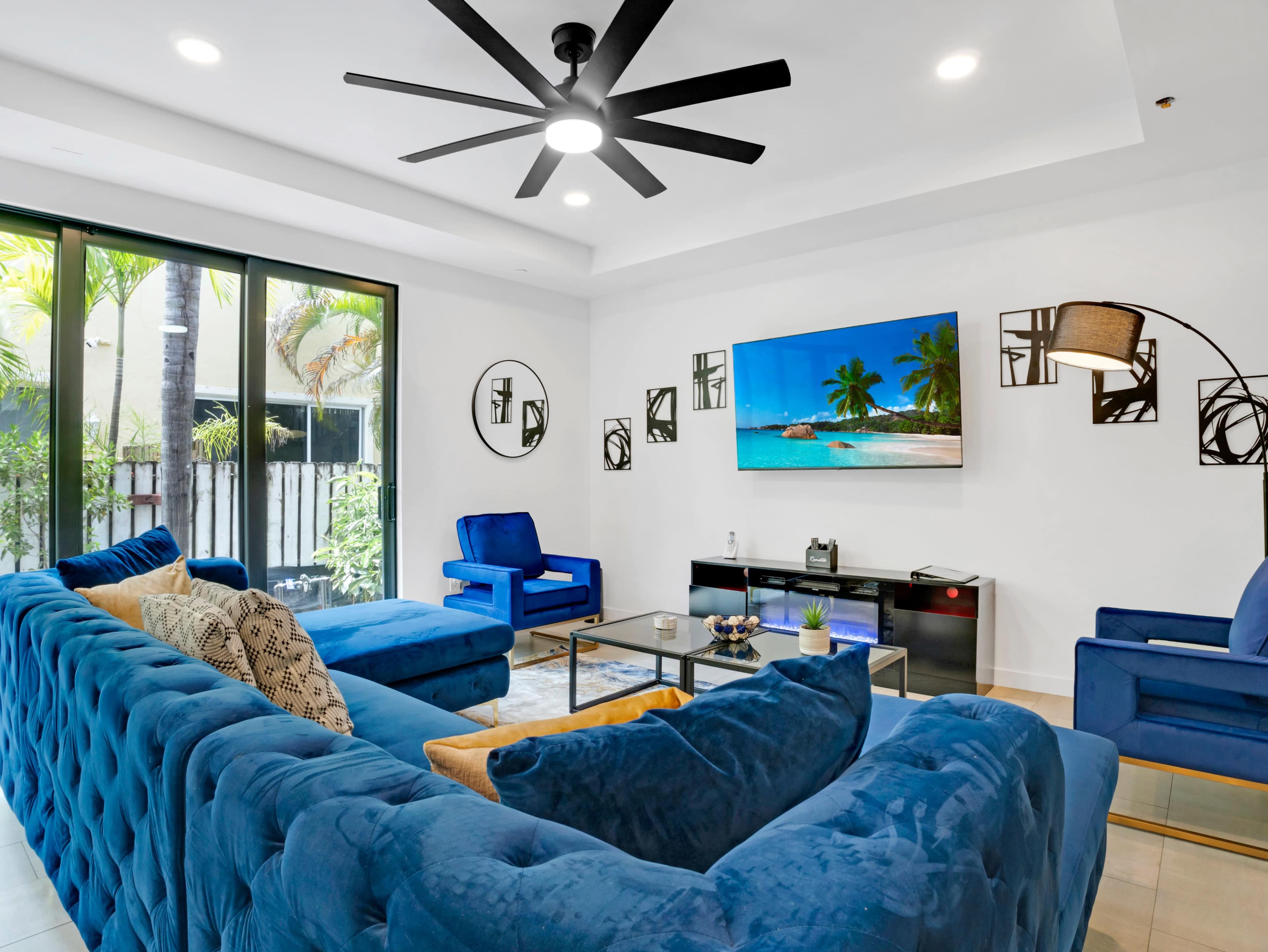 A spacious living room features a large blue sectional sofa, a wall-mounted television, and modern decor with palm trees visible through large windows.