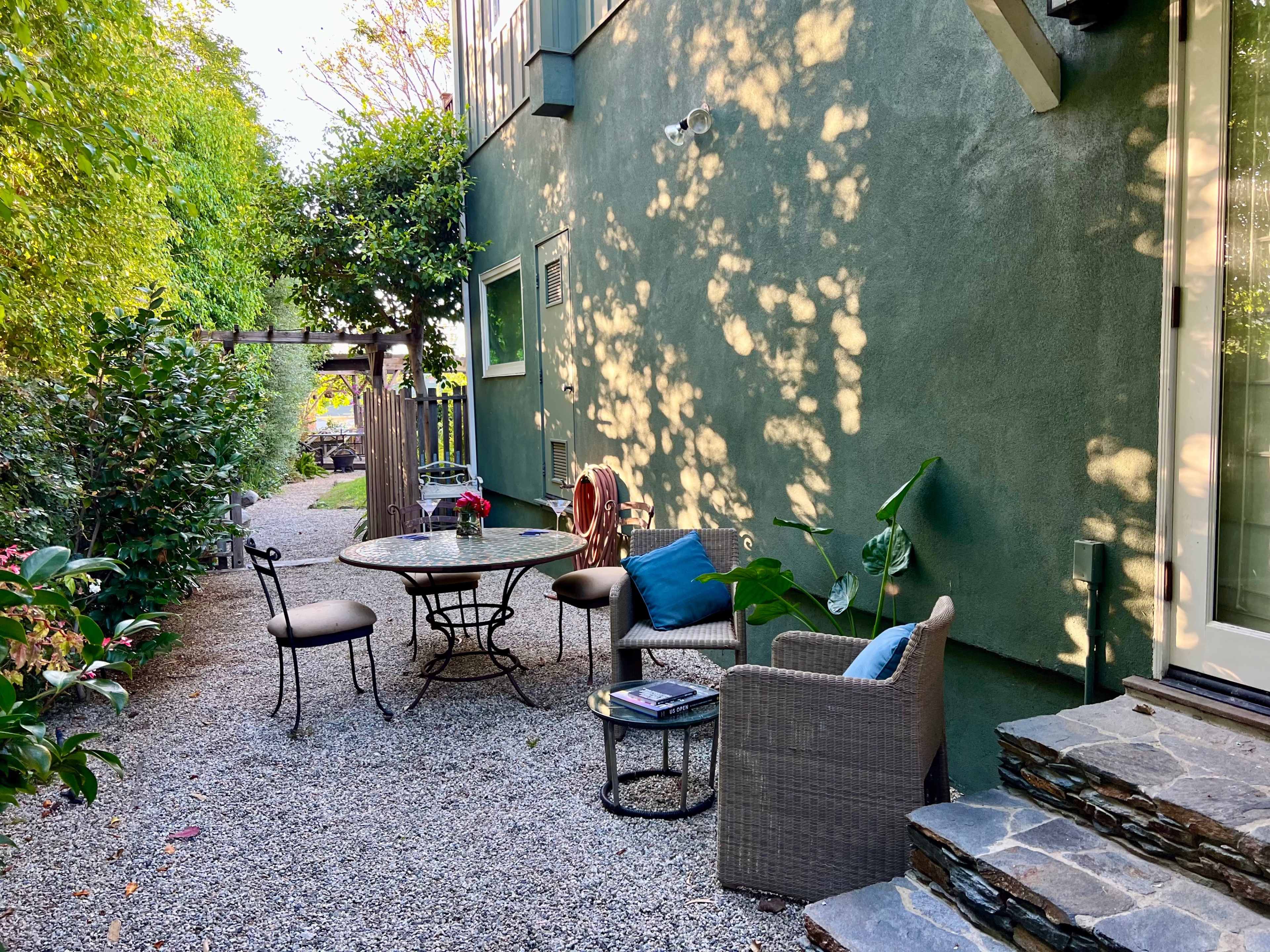 A small outdoor seating area is set on gravel beside a green house, featuring a table with chairs and surrounded by plants.