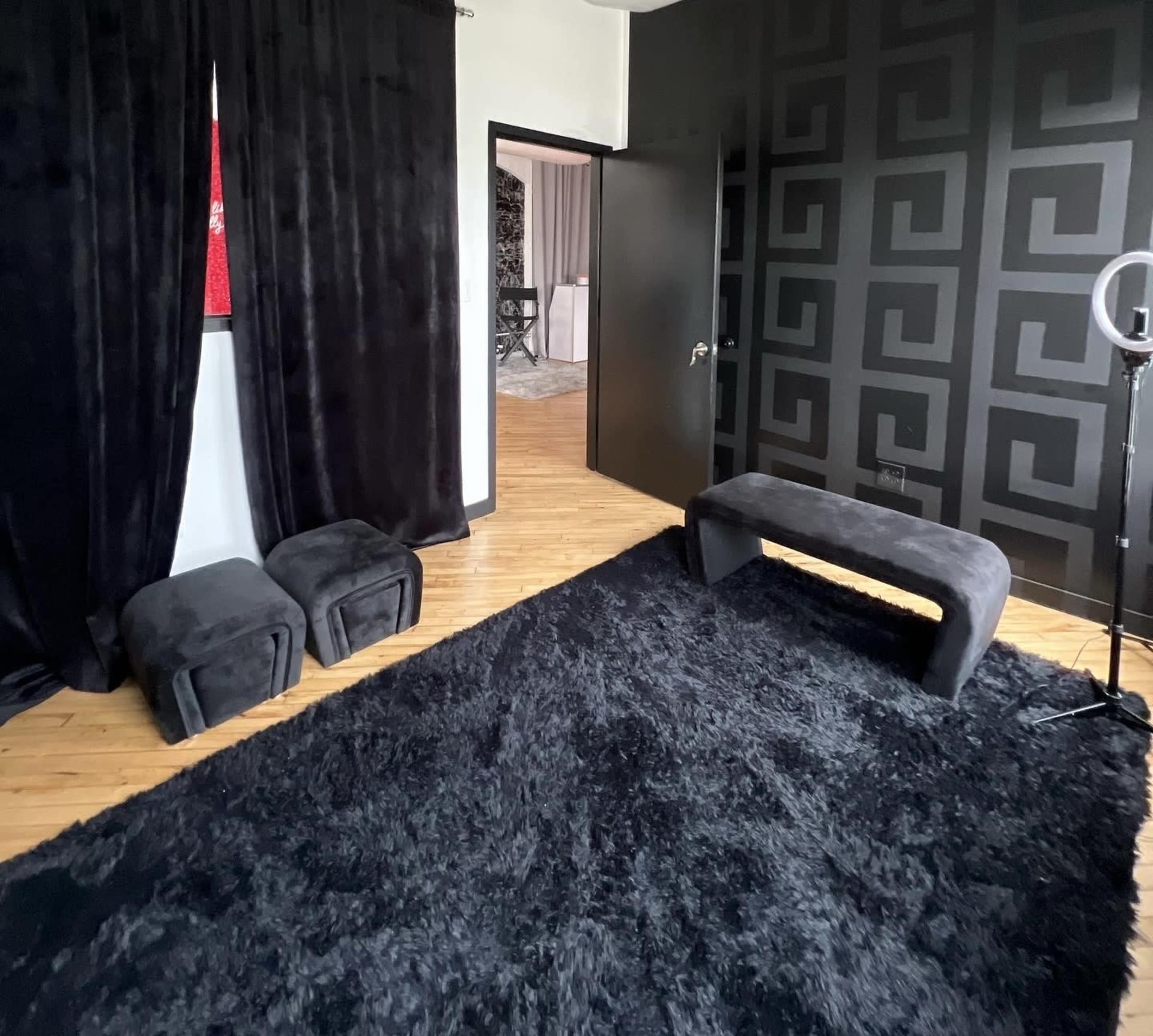 A room with black curtains, a dark patterned wall, a black rug, a low bench, and two small ottoman-style chairs.