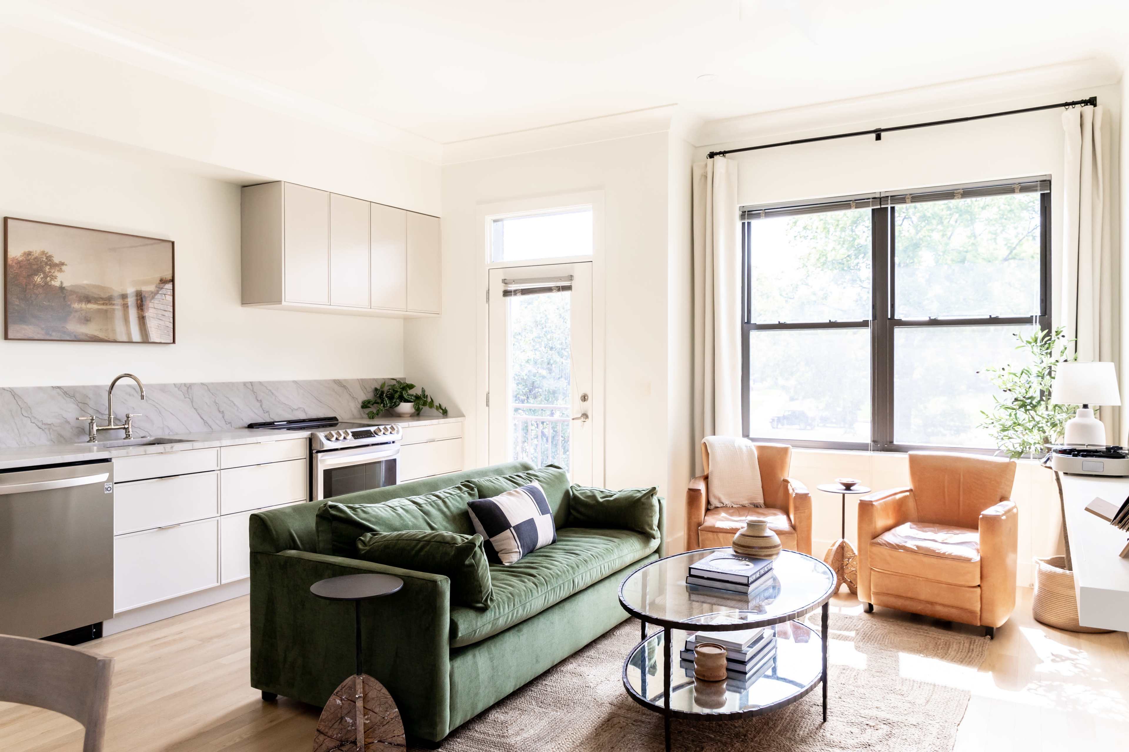 A modern living room features a green sofa, a round glass coffee table, and two orange armchairs, with a kitchen area visible in the background.
