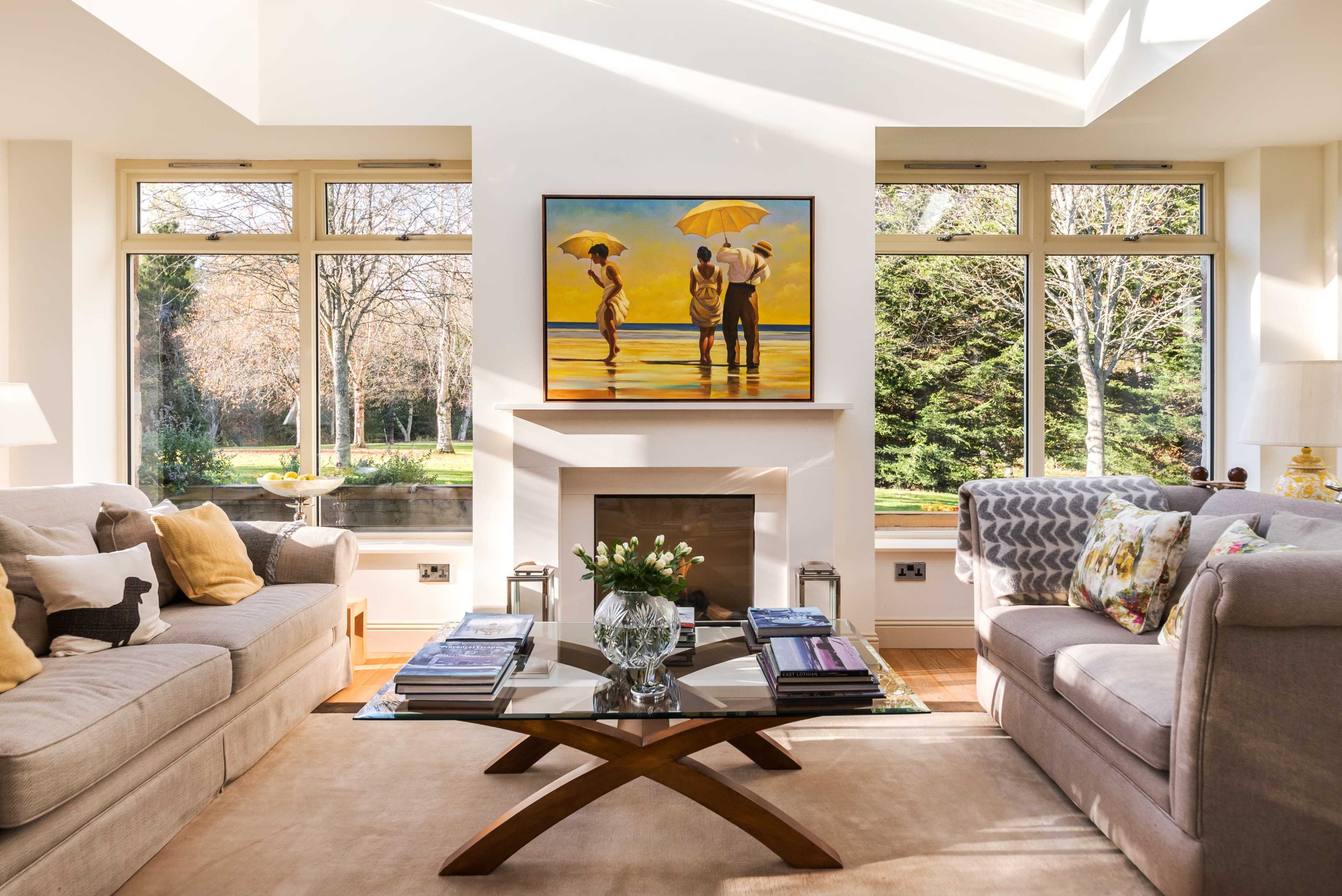 A bright living room with two sofas facing a coffee table and a large painting of three figures with umbrellas above a fireplace, flanked by large windows overlooking a garden.
