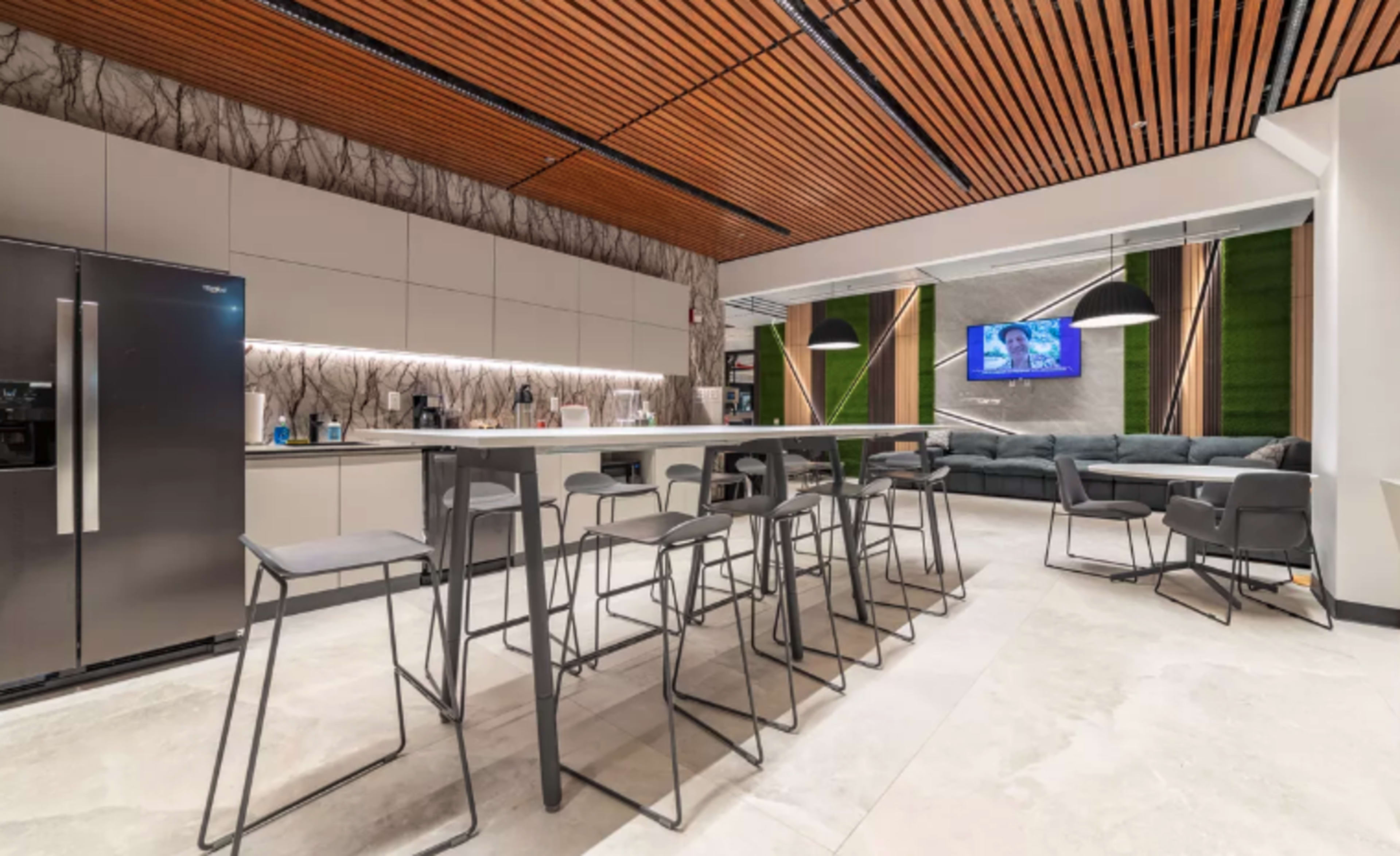 A modern kitchen and lounge area featuring a long bar with high stools, a refrigerator, and a seating area with a large couch and a television.
