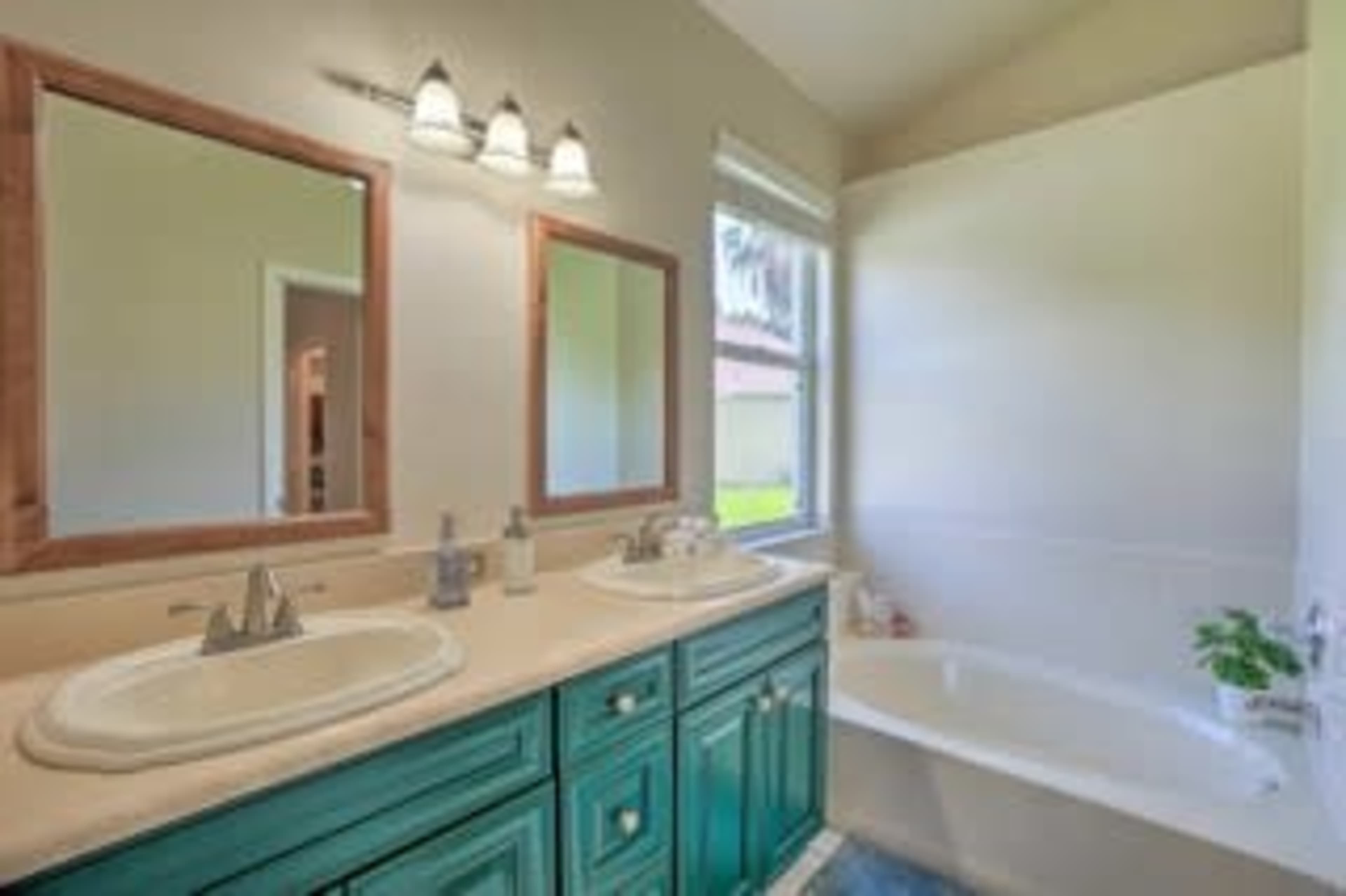 A bathroom featuring a double vanity with green cabinets, two mirrors, and a bathtub next to a window.