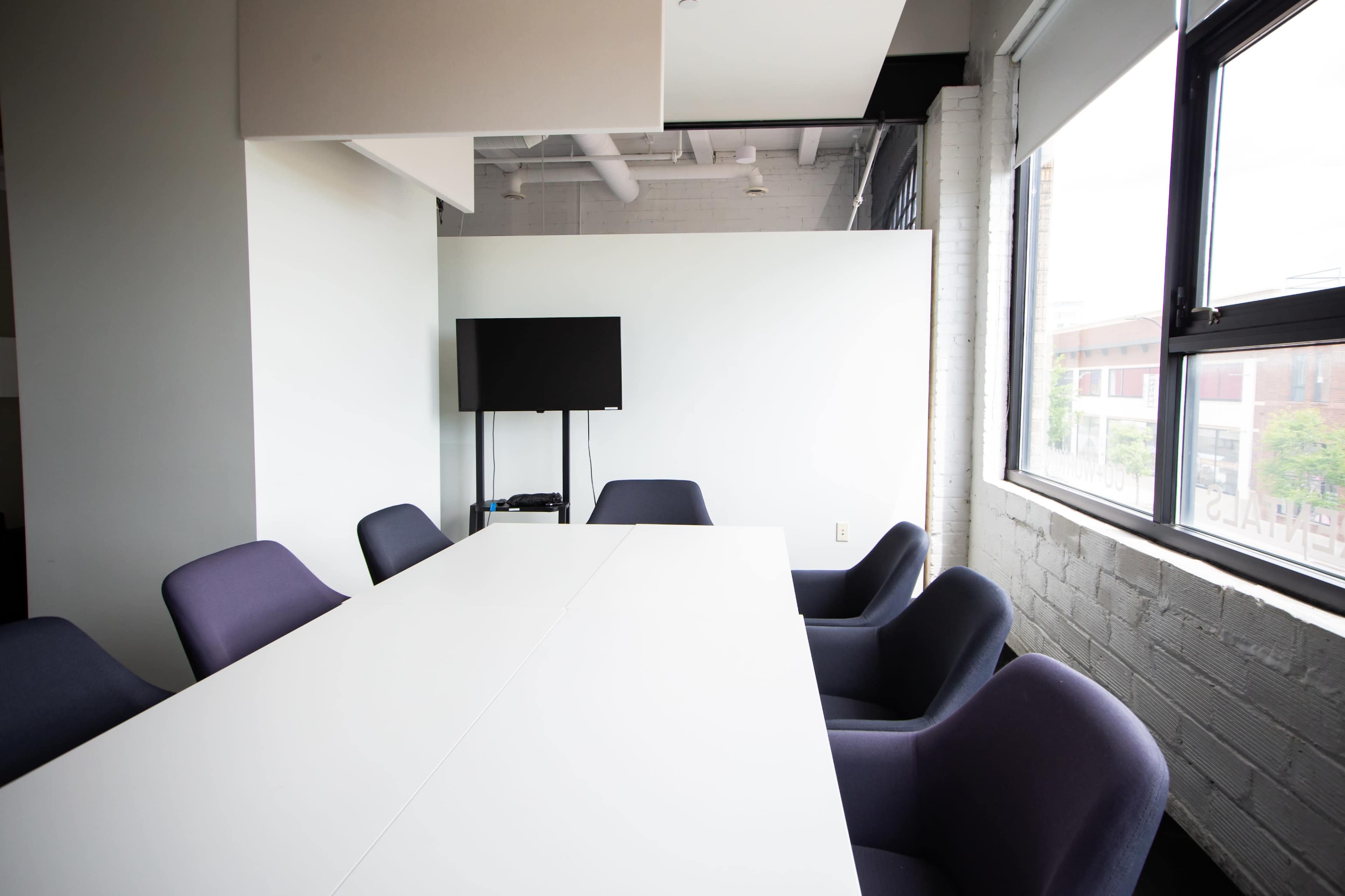 The image shows a modern conference room with a long white table surrounded by purple chairs and a television mounted on a wall.