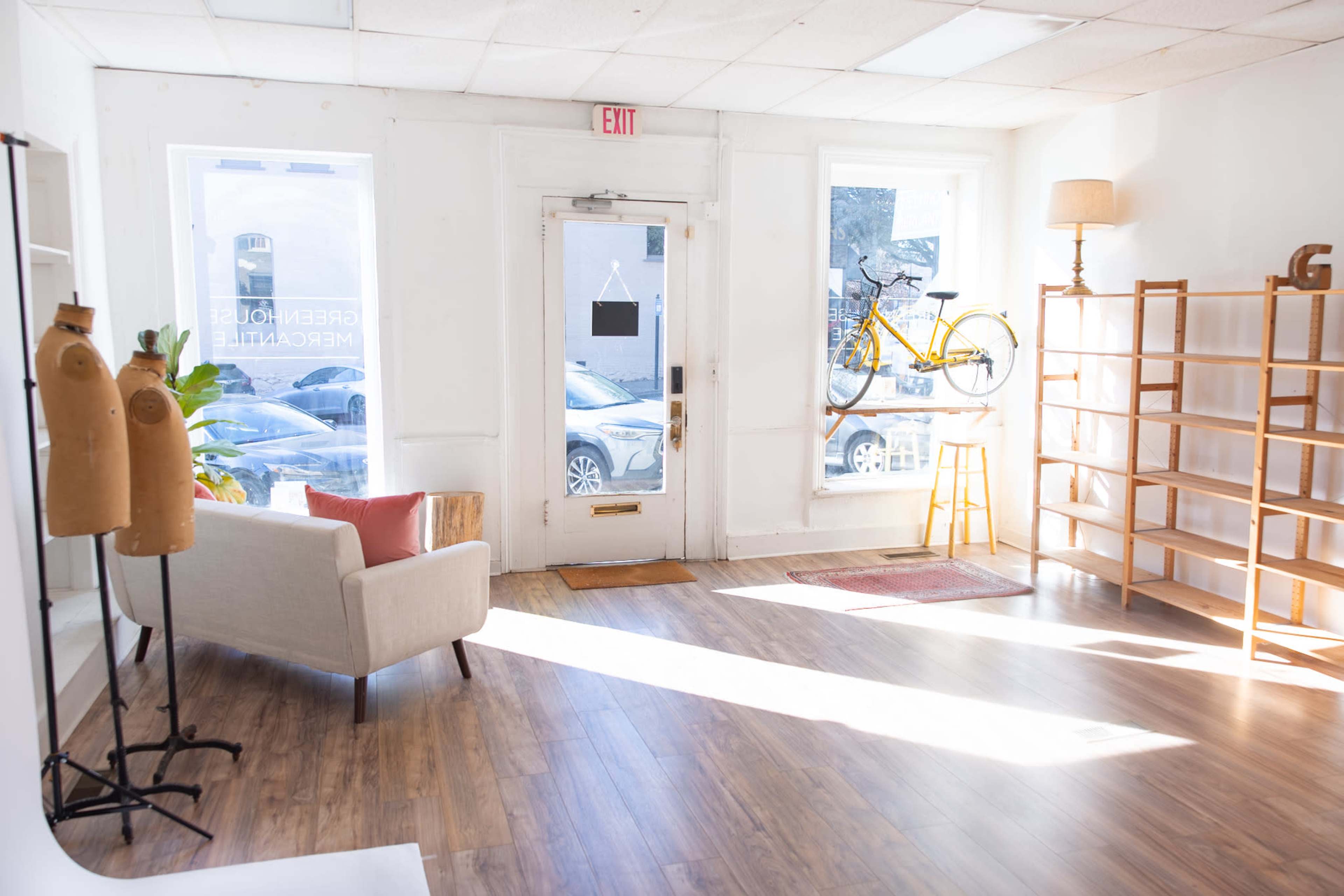 The image shows a bright, airy room with a bicycle mounted above the entrance, a comfortable white sofa on one side, and a large shelving unit on the other.