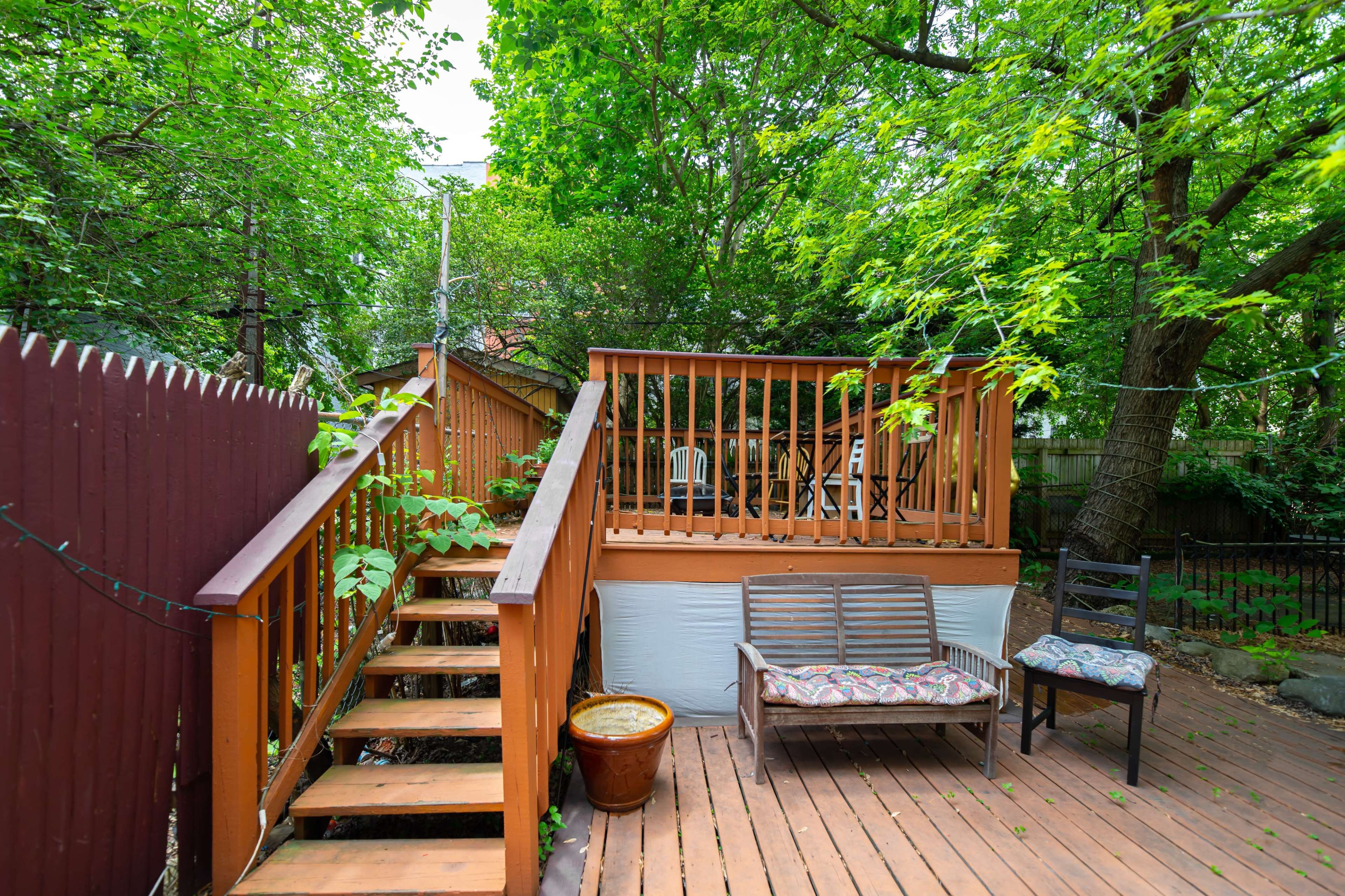 A wooden deck with chairs and a staircase leads to a patio surrounded by lush green trees.