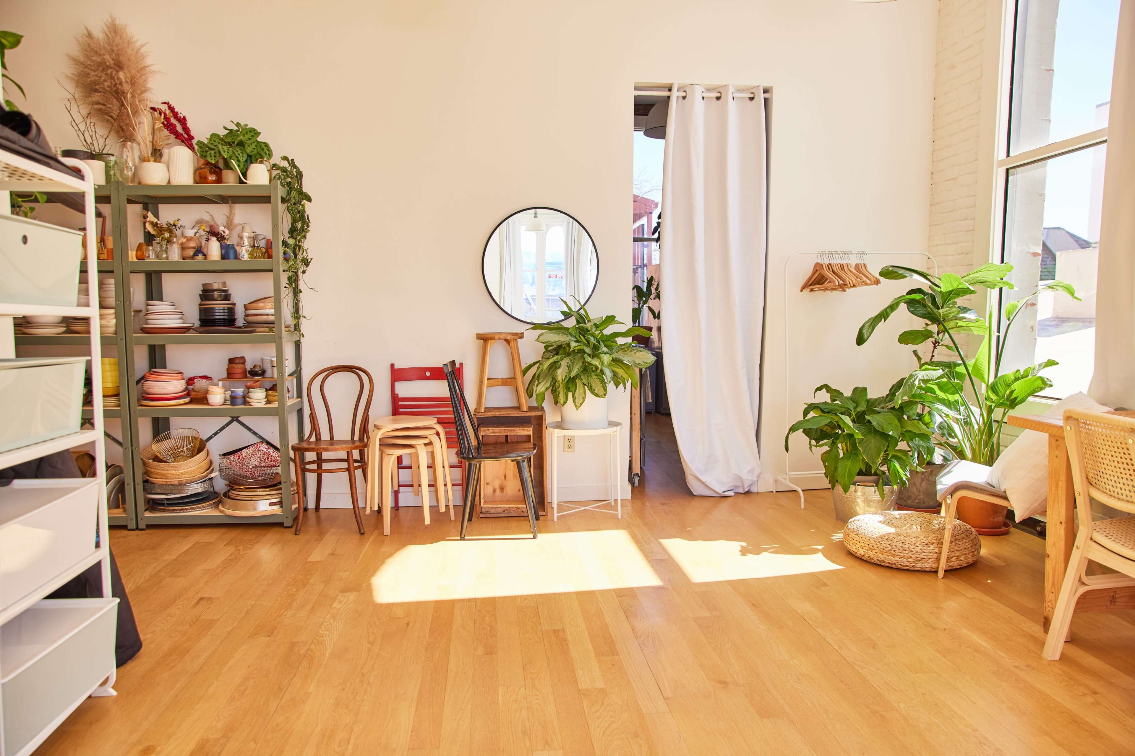 A well-lit room with light wooden floors, featuring a shelf filled with various items, a mirror, plants, and a curtain-draped doorway leading to another space.