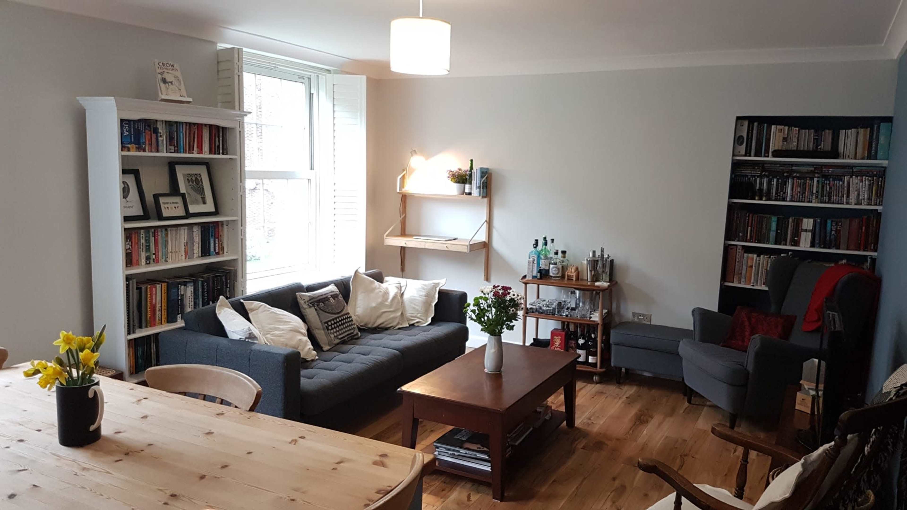 The image shows a cozy living room with a sofa, bookshelves filled with books, a wooden coffee table, and a dining area featuring a wooden table and chairs.