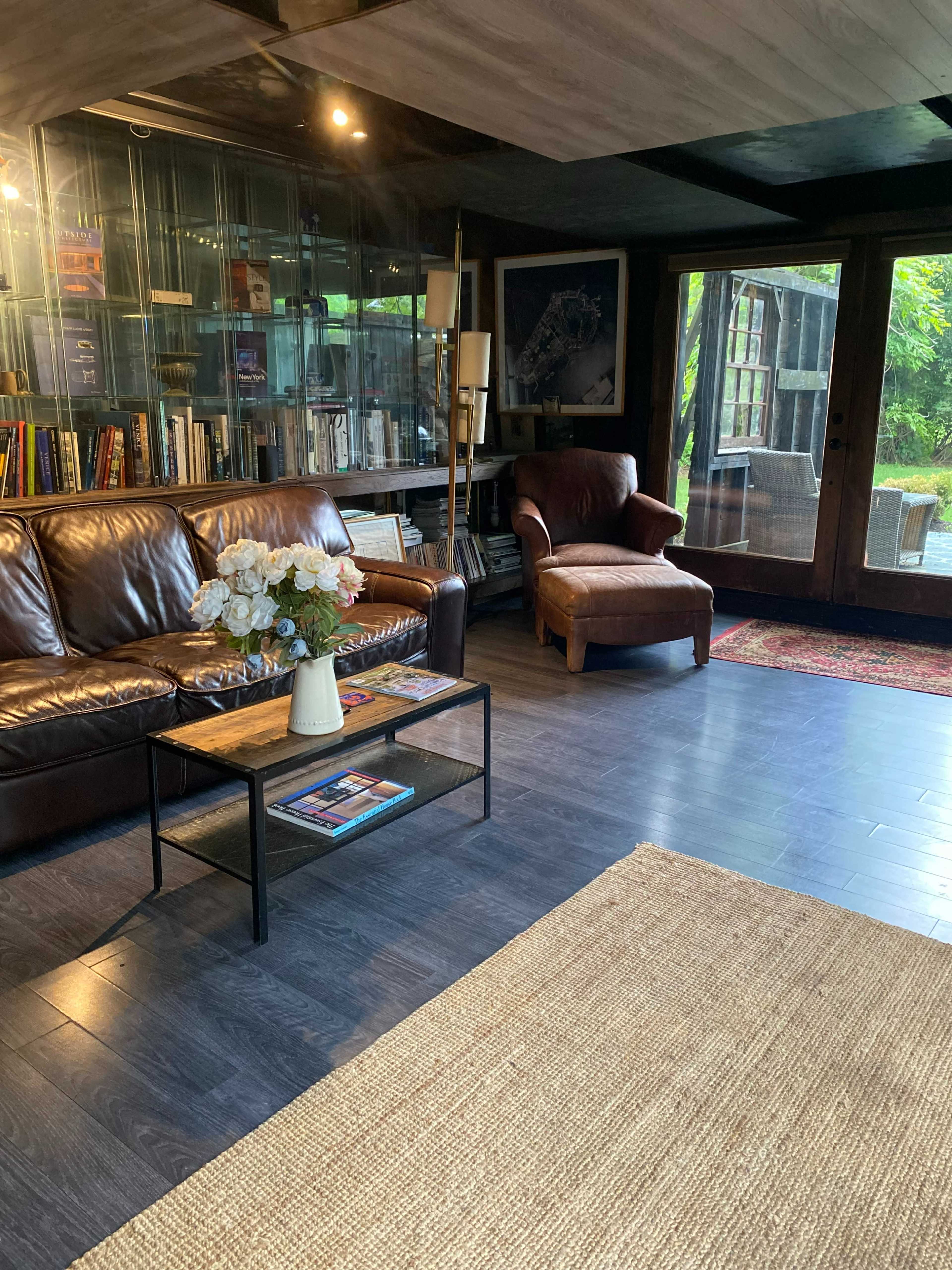 A cozy living room features a brown leather sofa, a wooden coffee table with magazines, a large bookcase, and a chair, with glass doors leading to an outdoor view.