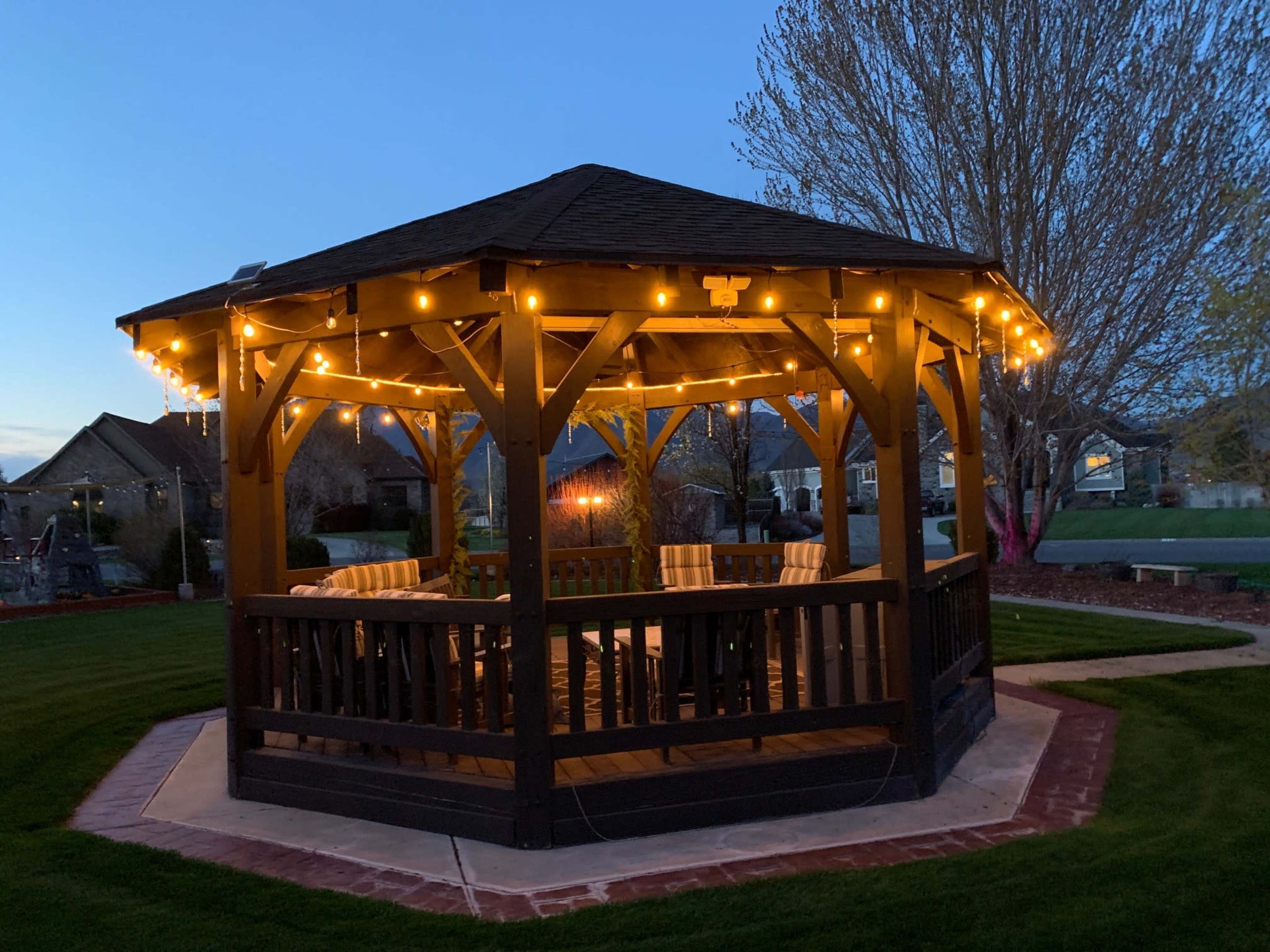 A wooden gazebo with string lights is illuminated in a landscaped yard during dusk.