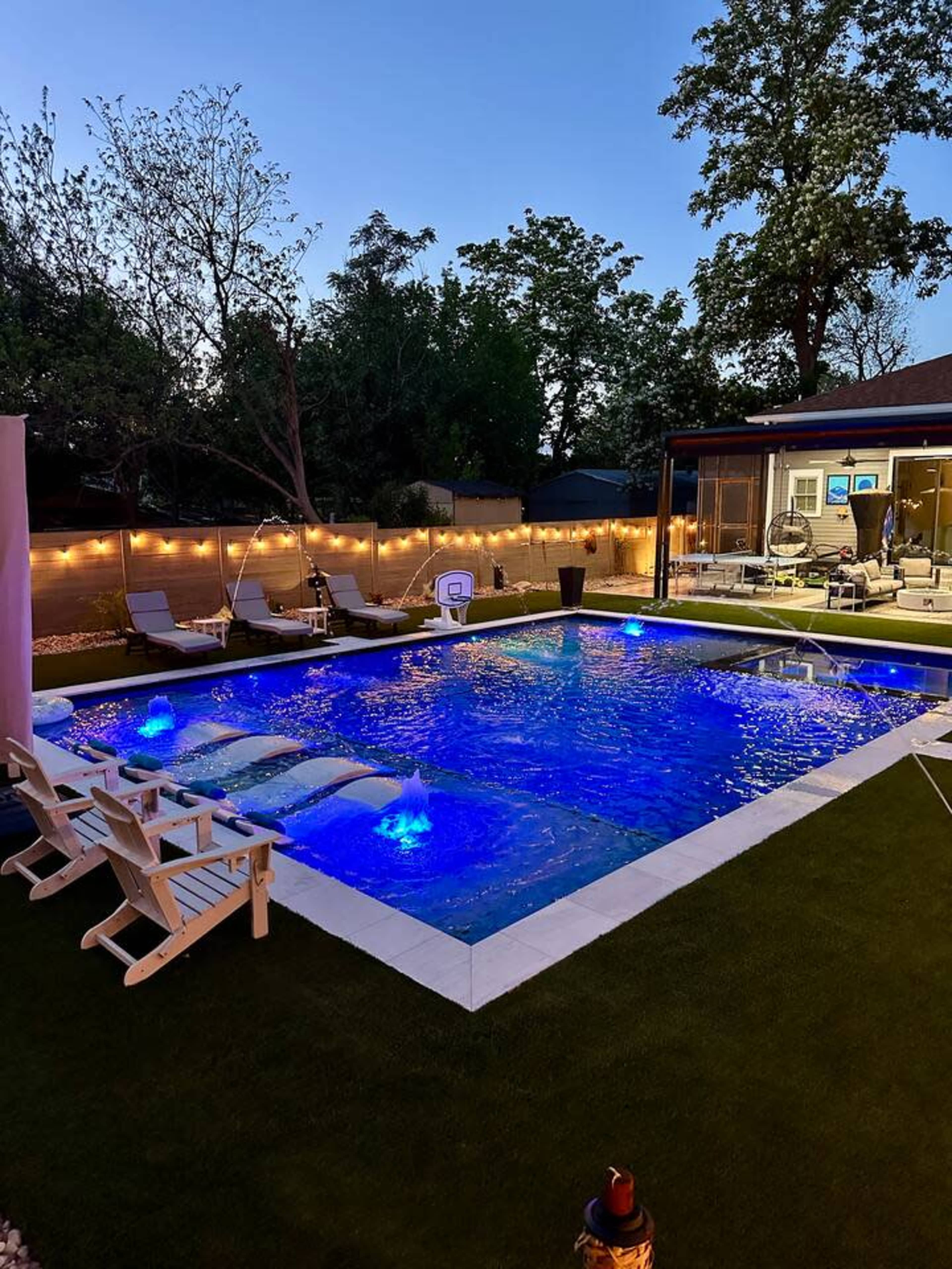 A rectangular pool is illuminated by blue lights, surrounded by lounge chairs and grass, with a modern house visible in the background under twilight.