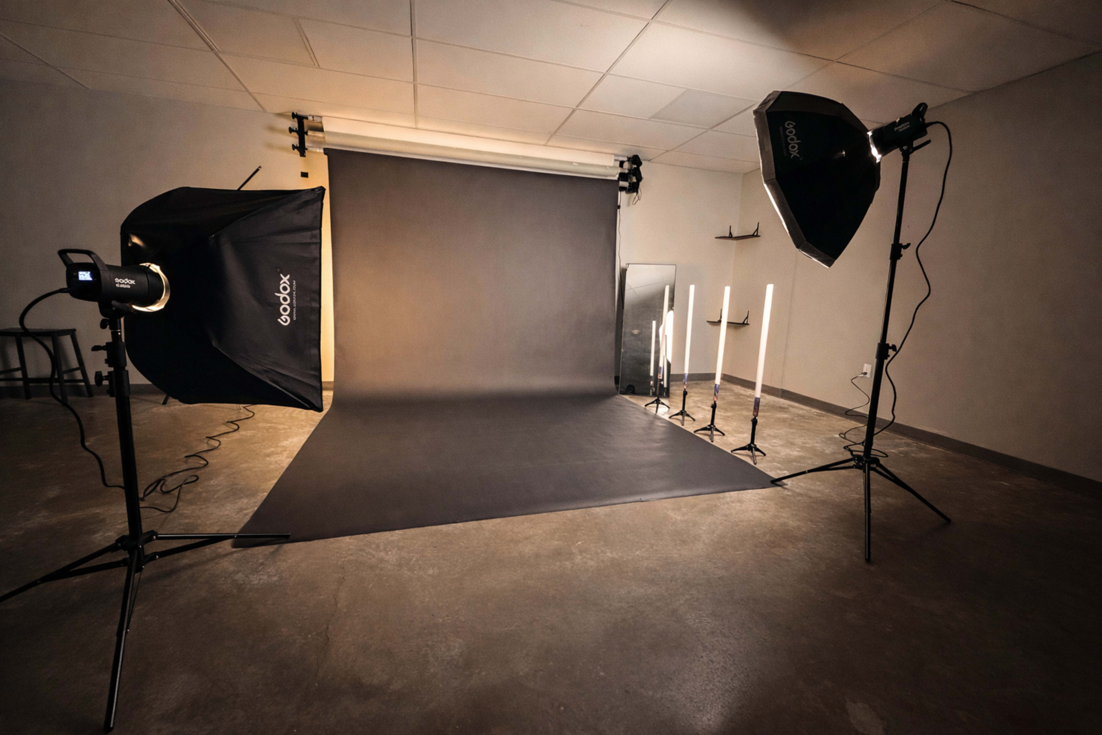 The image shows a photography studio setup with softbox lights, a backdrop, and various light stands positioned around a dark seamless background.