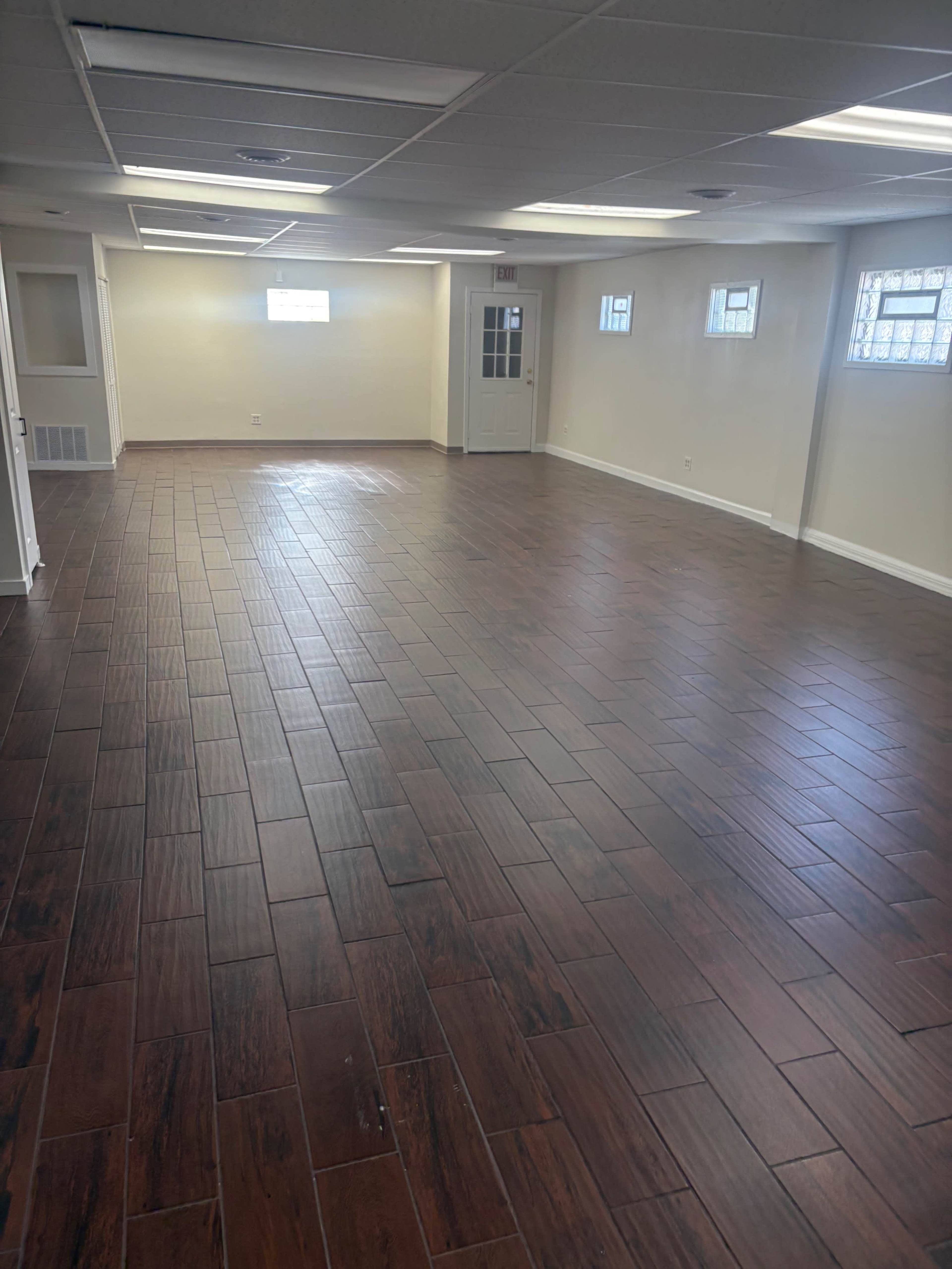 The image shows a spacious, empty room with brown tiled flooring and windows providing natural light.