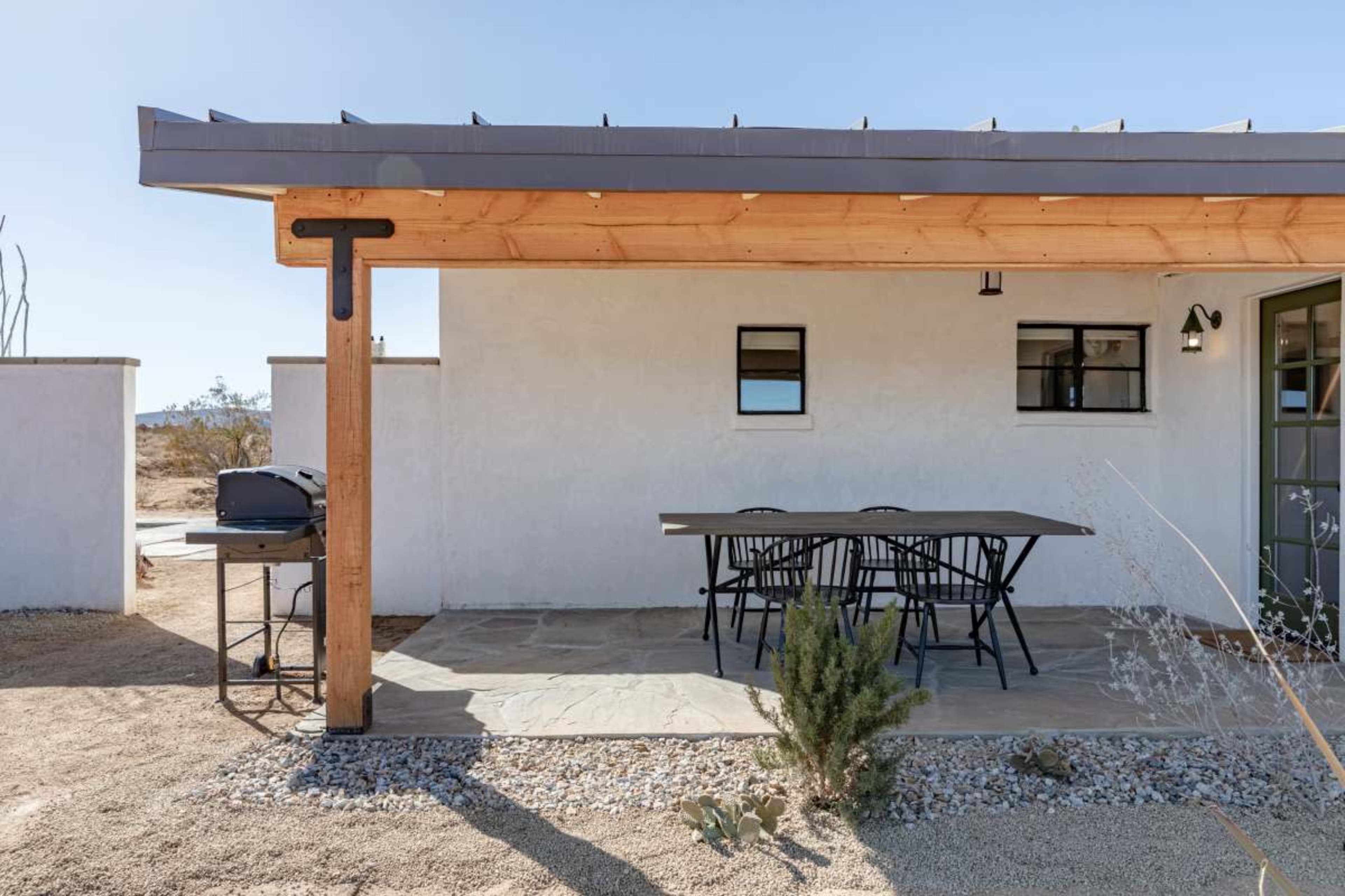 The image shows a small outdoor patio area with a black dining table and chairs, adjacent to a white building under a wooden overhang.